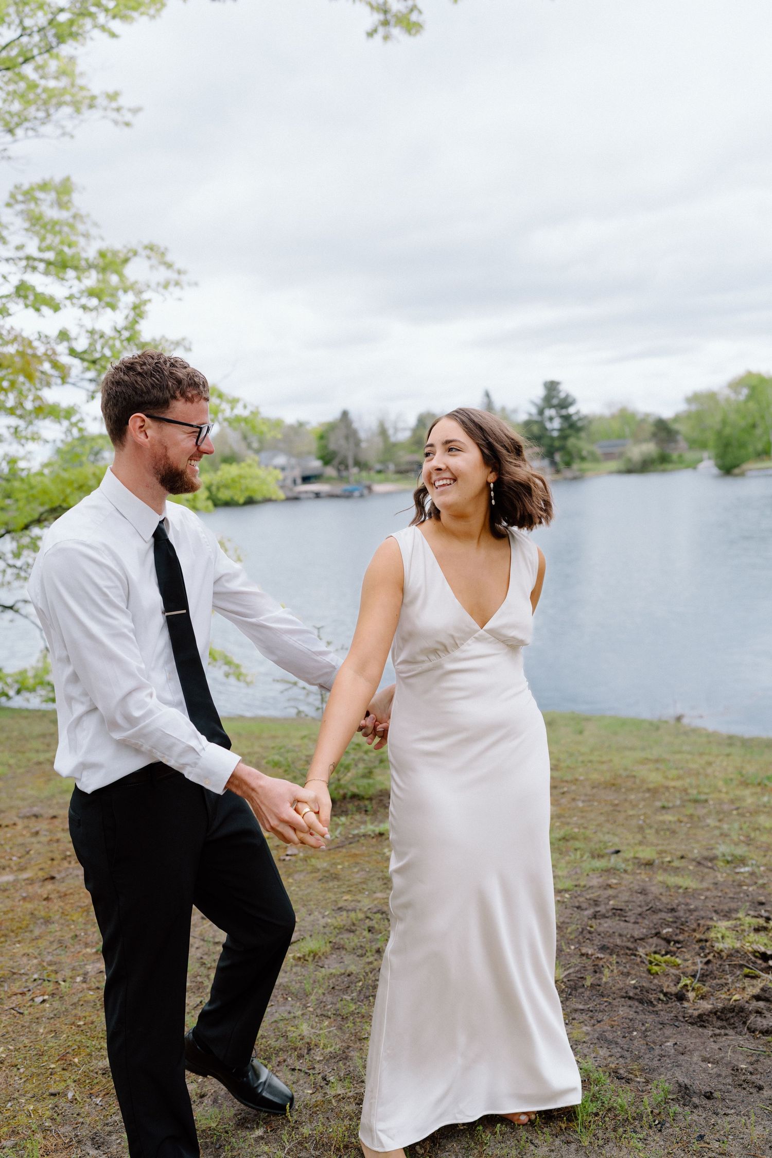 A couple dances and laughs together by a lake on an overcast day.