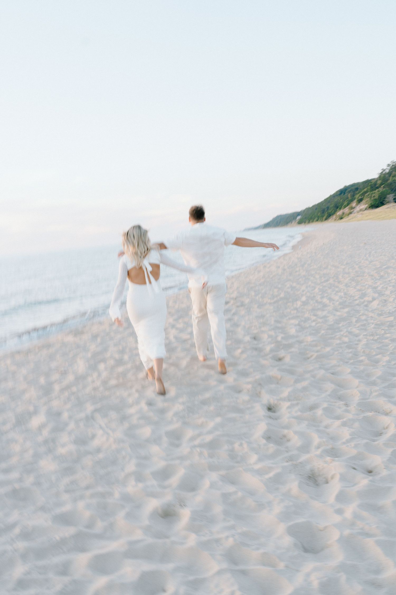 A couple runs together on a sandy beach wearing matching white outfits.