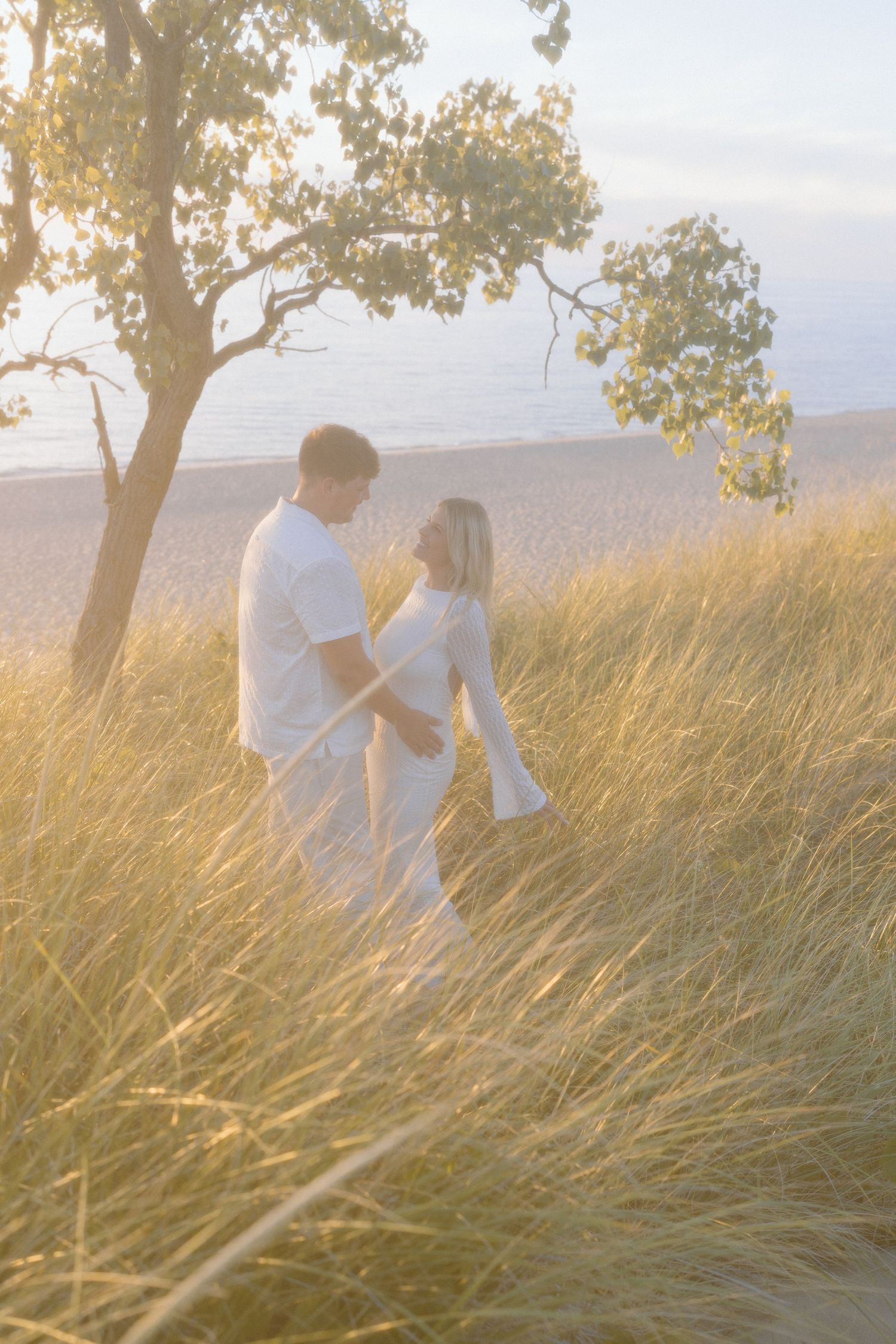 A romantic couple stands among tall grass at sunset near the ocean.