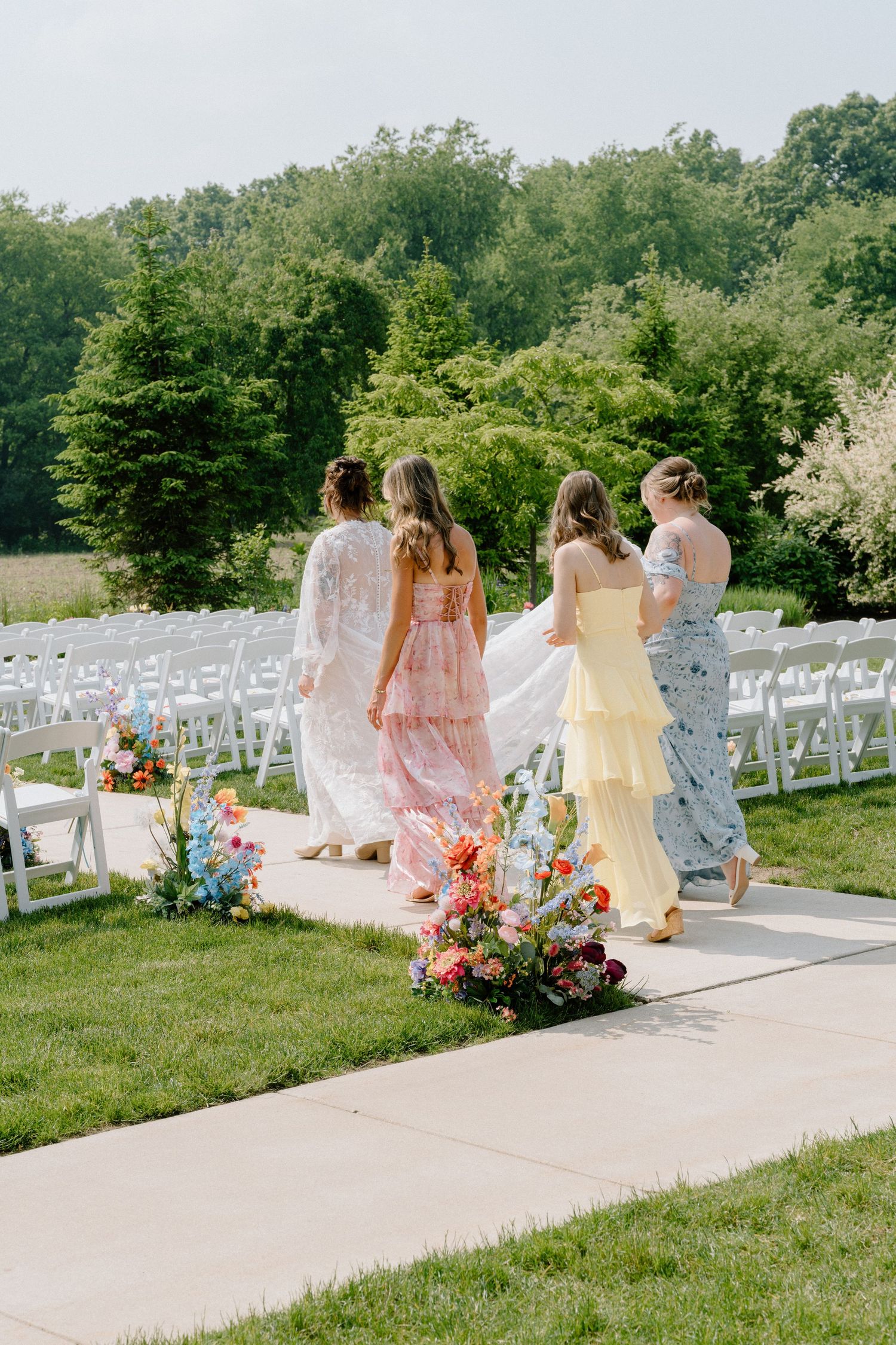 Wedding guests in pastel dresses walk along pathway lined with white ceremony chairs.