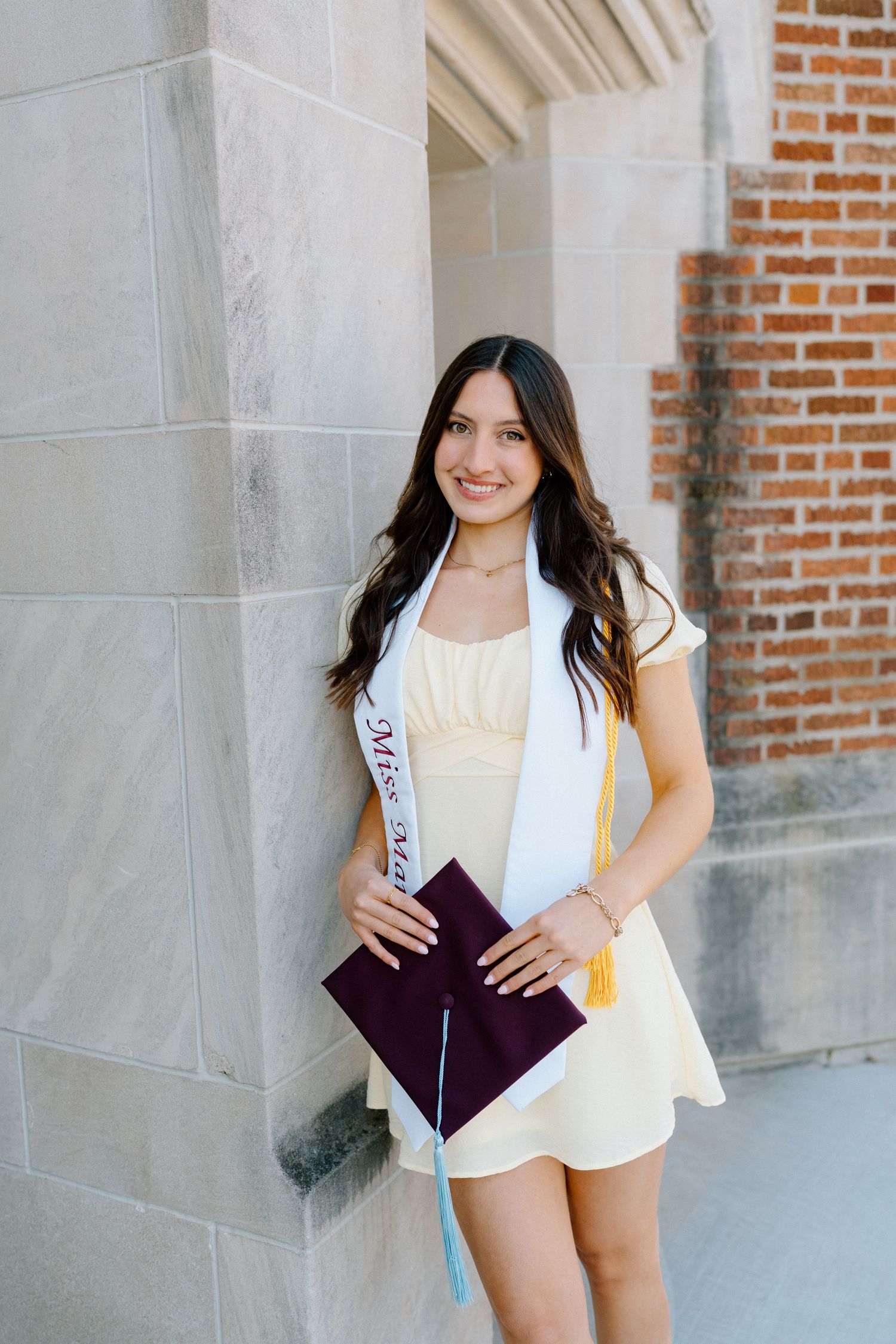 Smiling graduate in yellow dress holding maroon cap with gold tassel, wearing a Miss pageant sash outdoors.