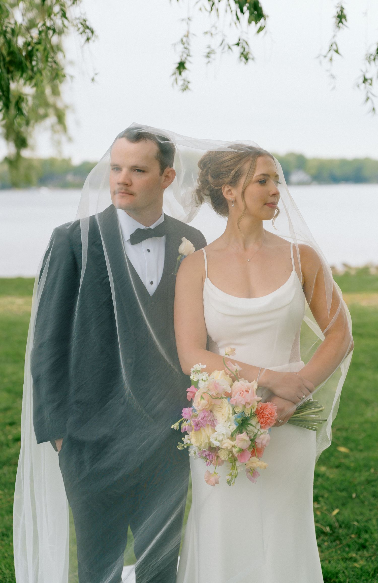 Bride in white gown with veil and groom in gray tuxedo pose outdoors near a lake holding a colorful bouquet.