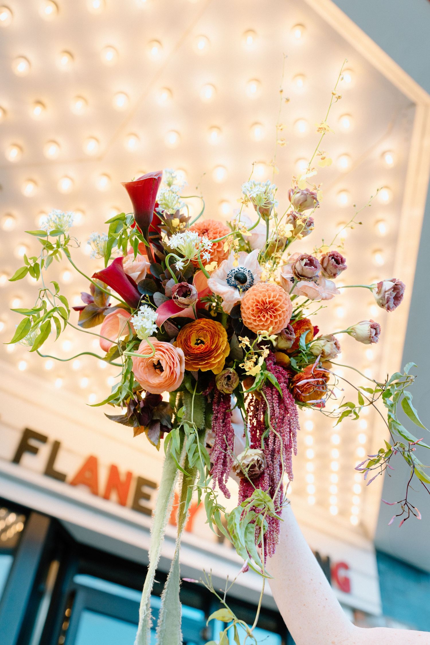 Elegant floral arrangement with pink and burgundy blooms in tall white vase against string lights and blue wall decor.