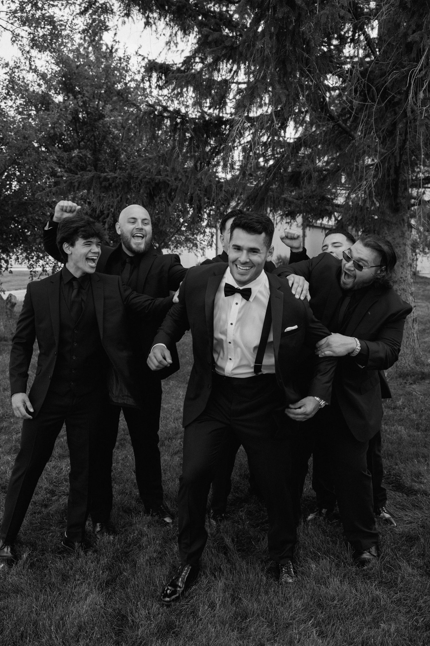 Groomsmen in black suits pose playfully on grass in black and white photography.