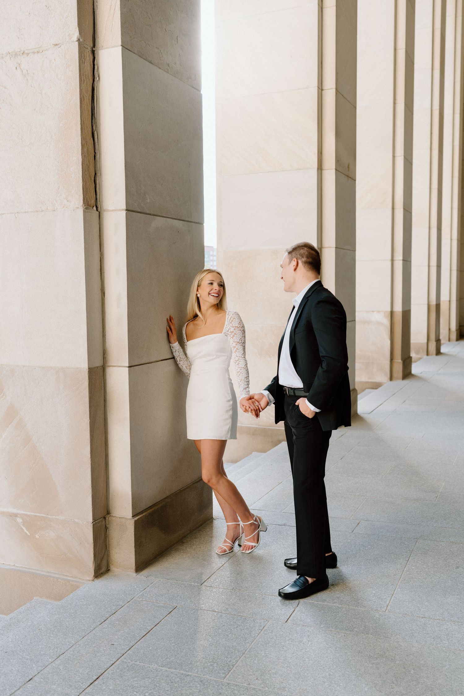 A couple shares a playful moment in an elegant colonnade hallway with white stone columns and grey floors.