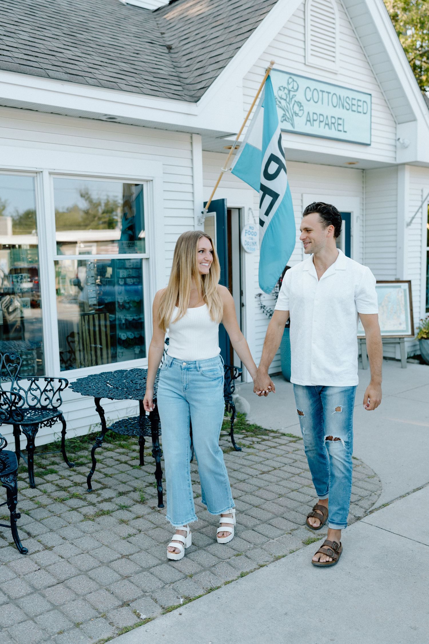 A couple walks hand in hand outside a white cottage shop with teal flags.