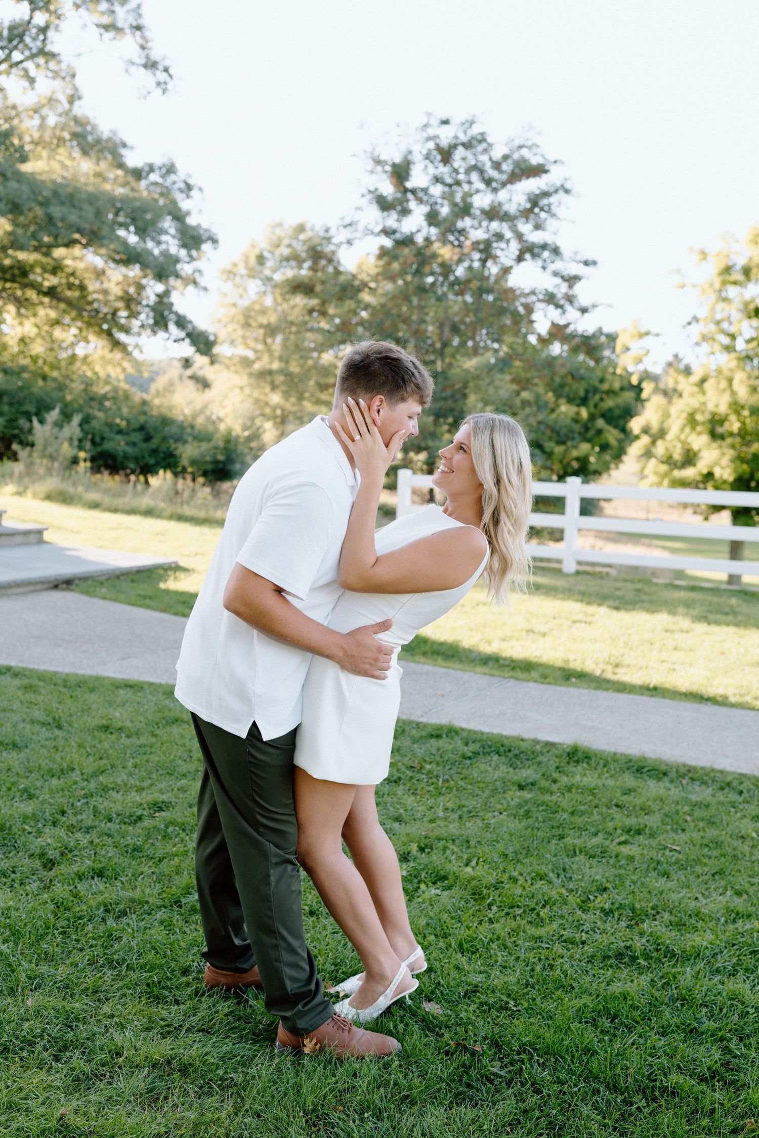 A playful engagement photoshoot in a grassy park with white fence backdrop.