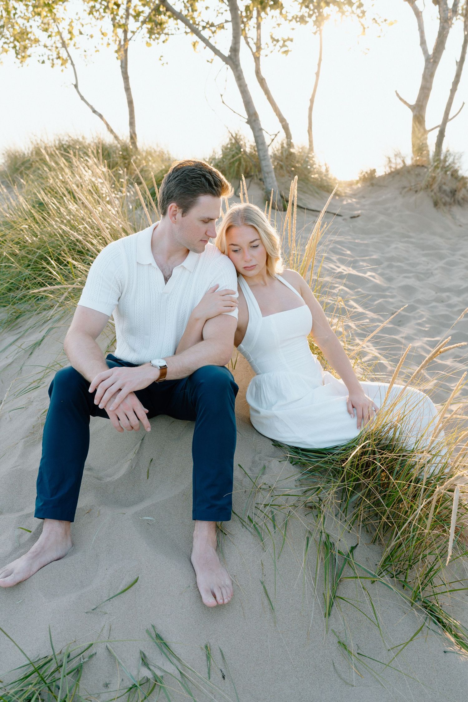 A couple sits together intimately on a sandy beach with beach grass at sunset.