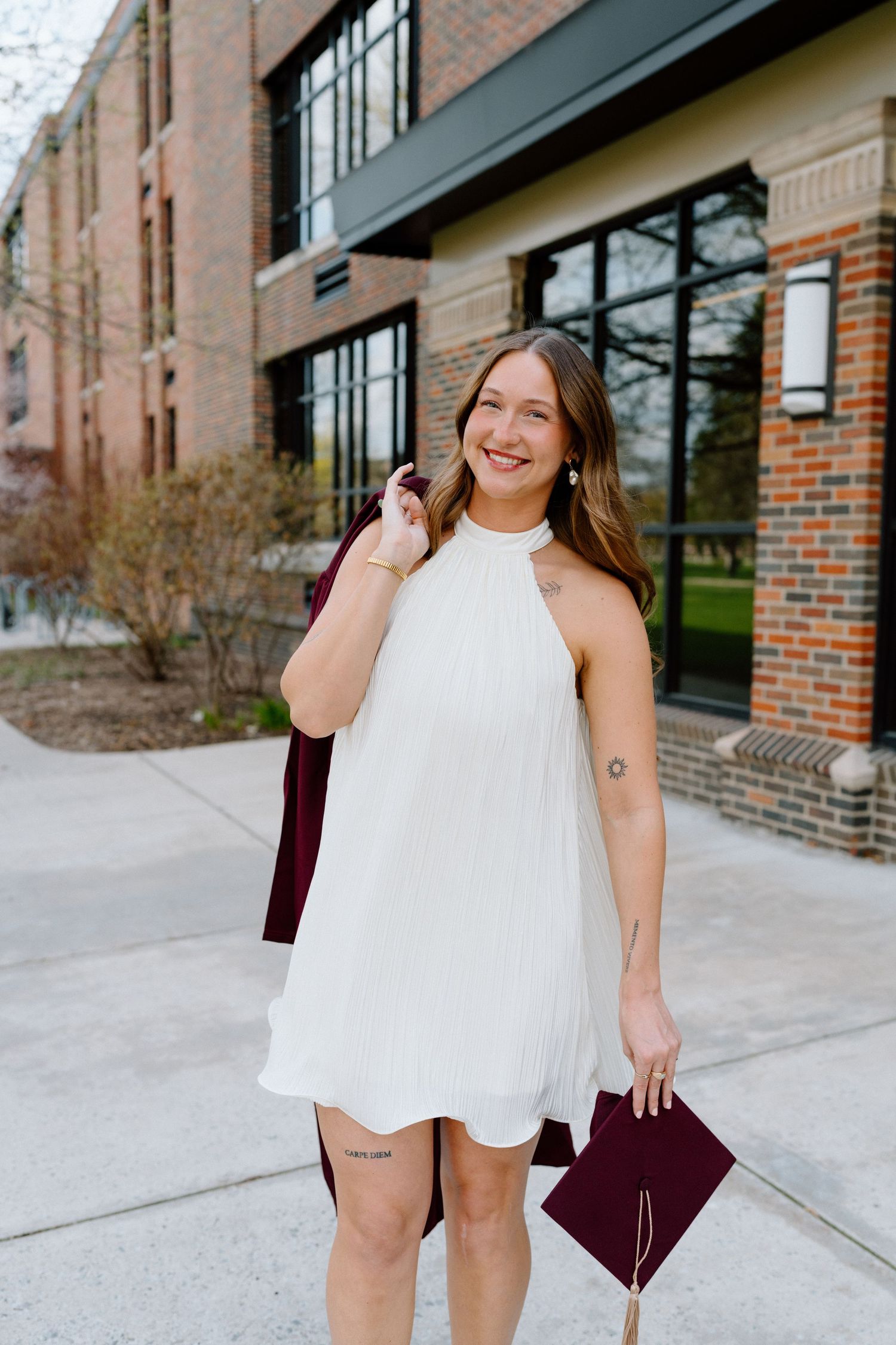 Smiling graduate in white halter dress holding maroon cap and gown outdoors near brick building.