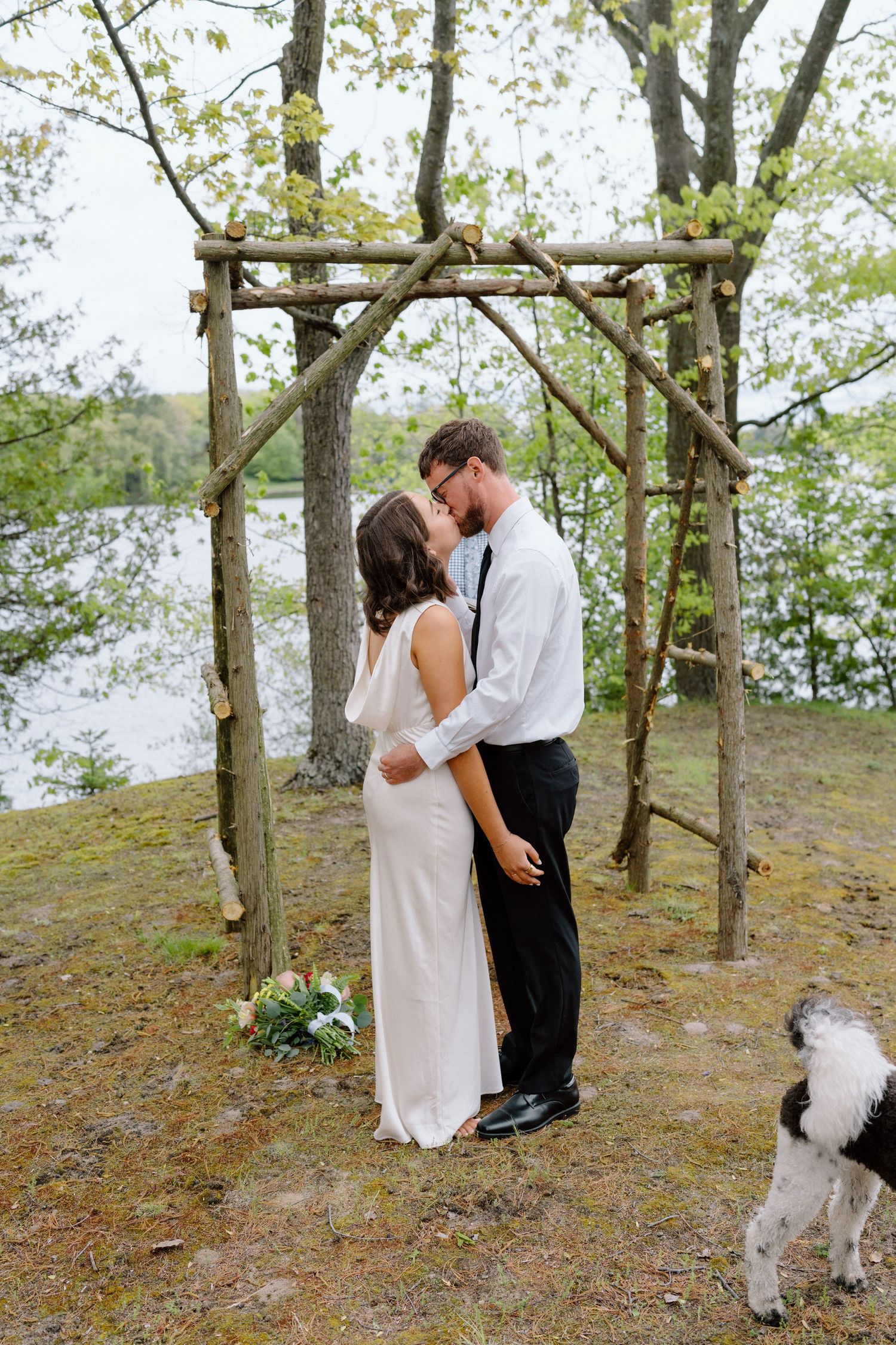 A couple shares an intimate moment under trees while their dog watches nearby.