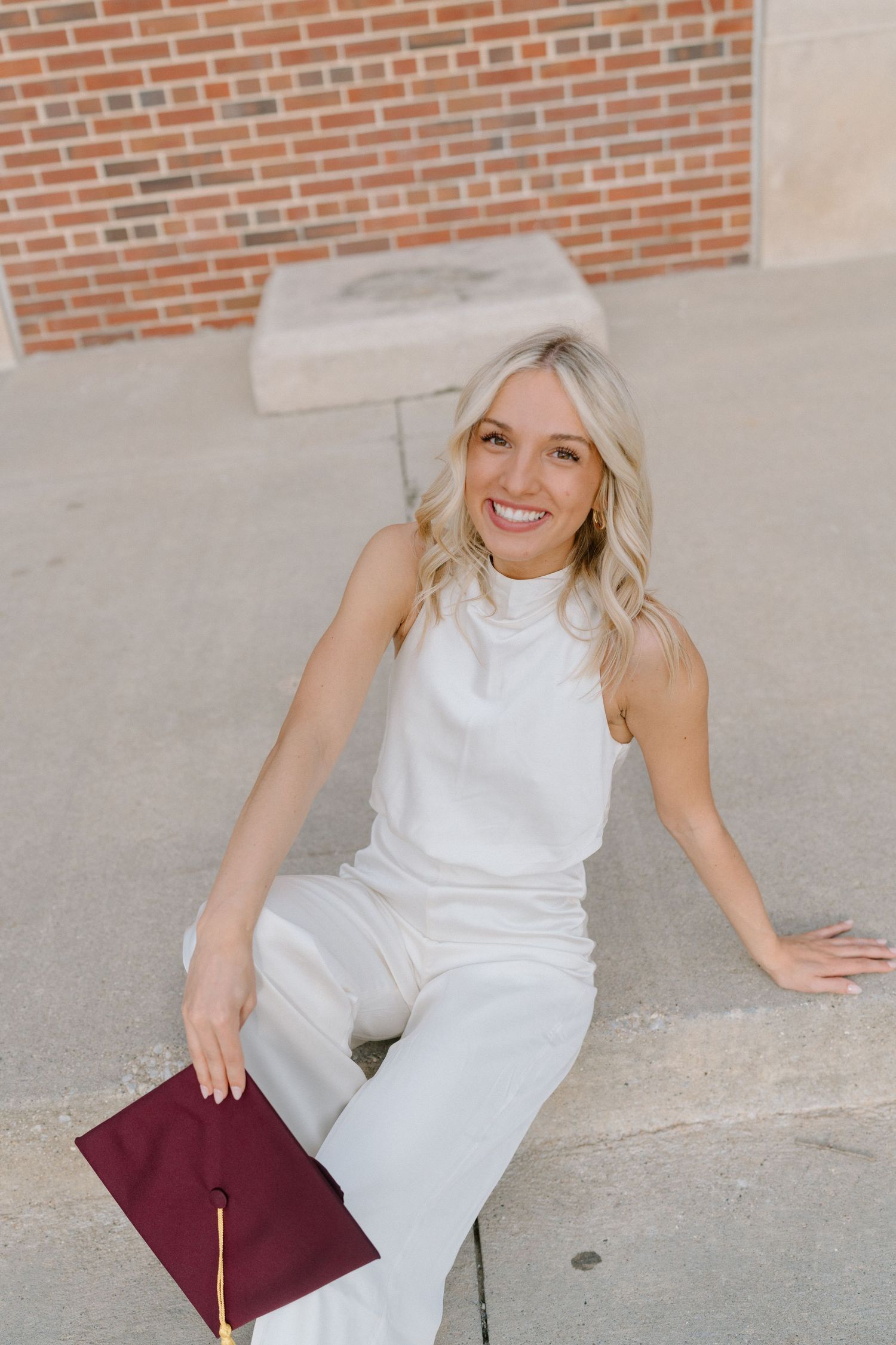 Graduate in white outfit sits casually with burgundy diploma on concrete steps.