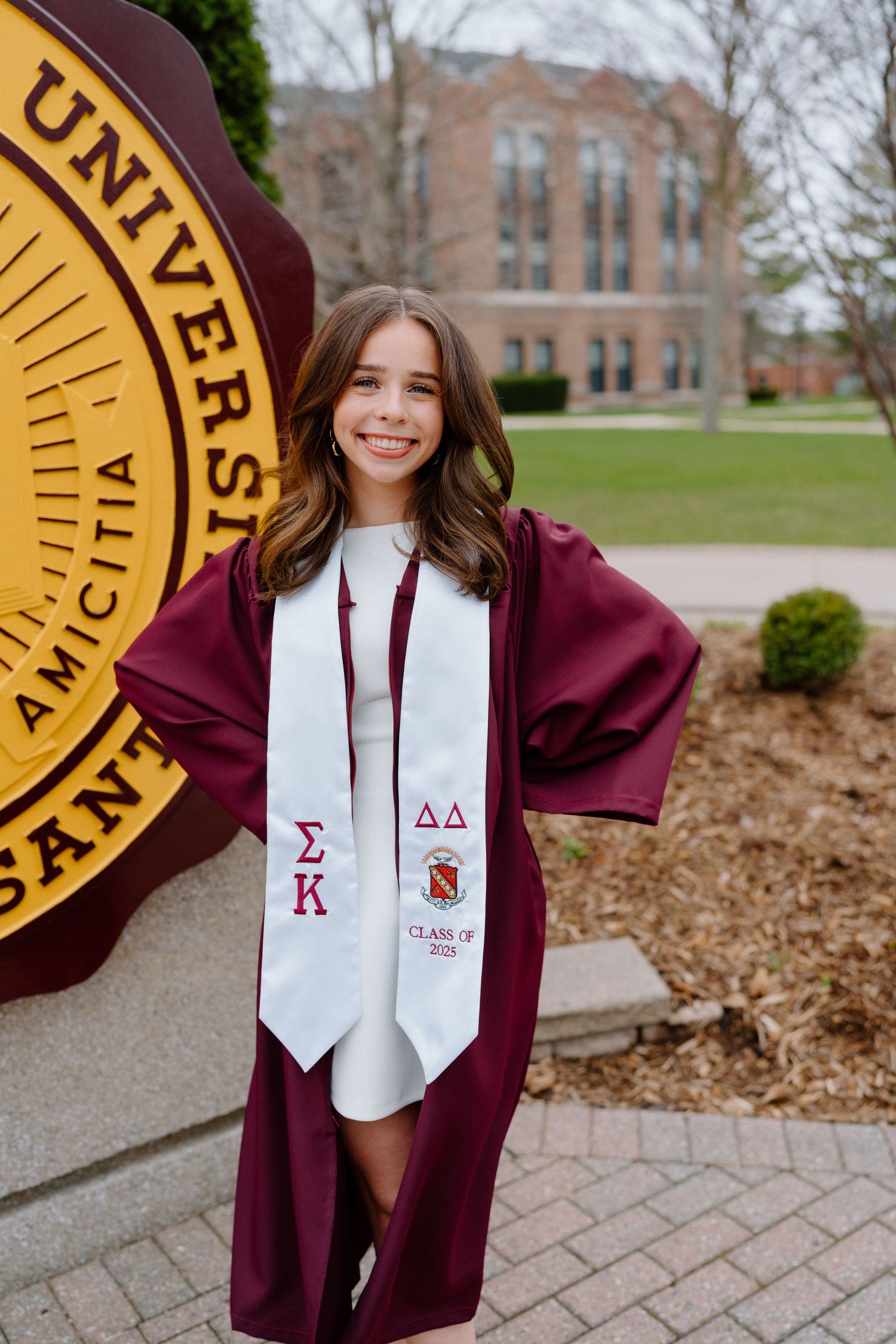Graduate in burgundy regalia displays sorority stole and honor cords by university seal.
