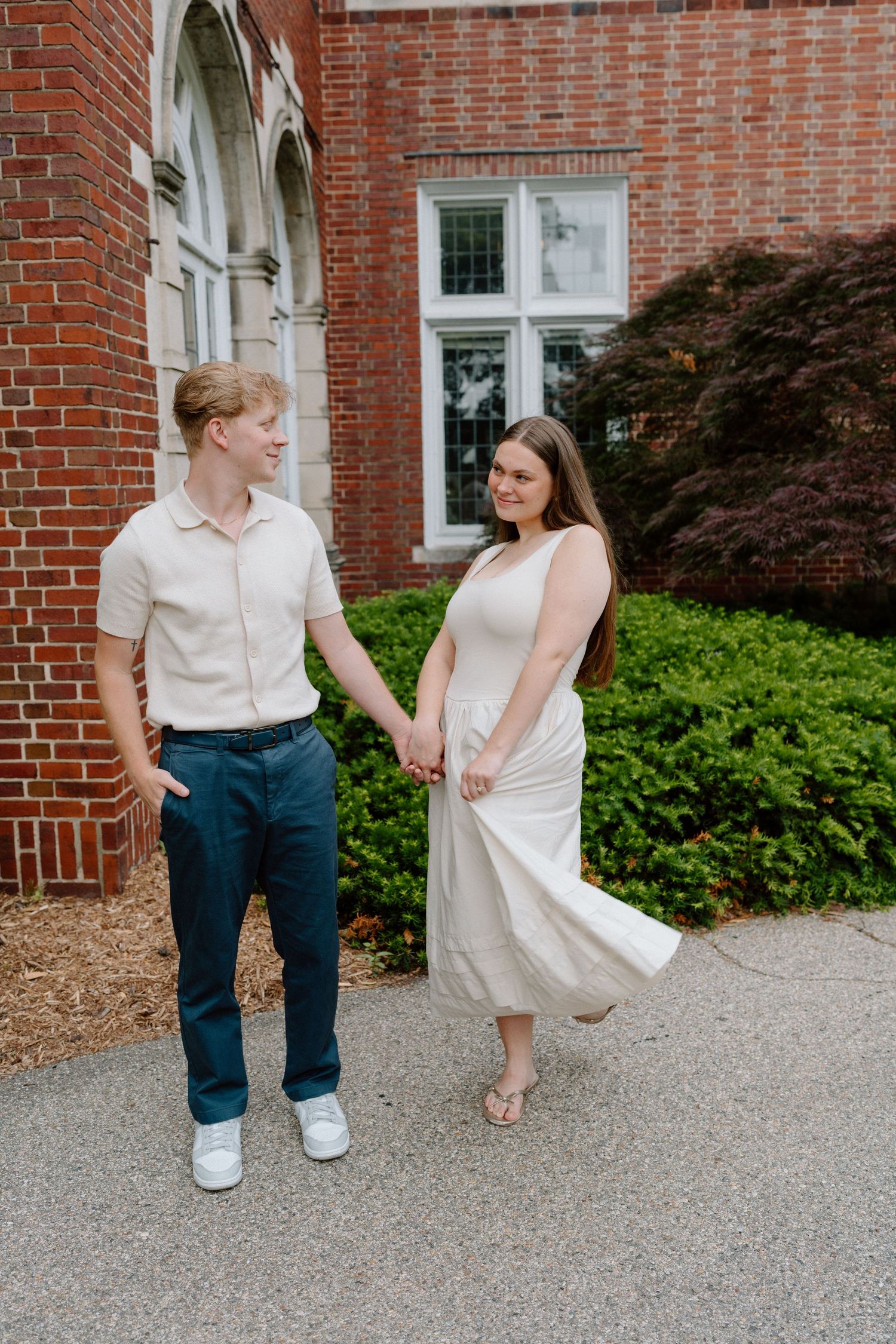 A couple in coordinated white and blue outfits walks together on a gravel path beside a brick building.