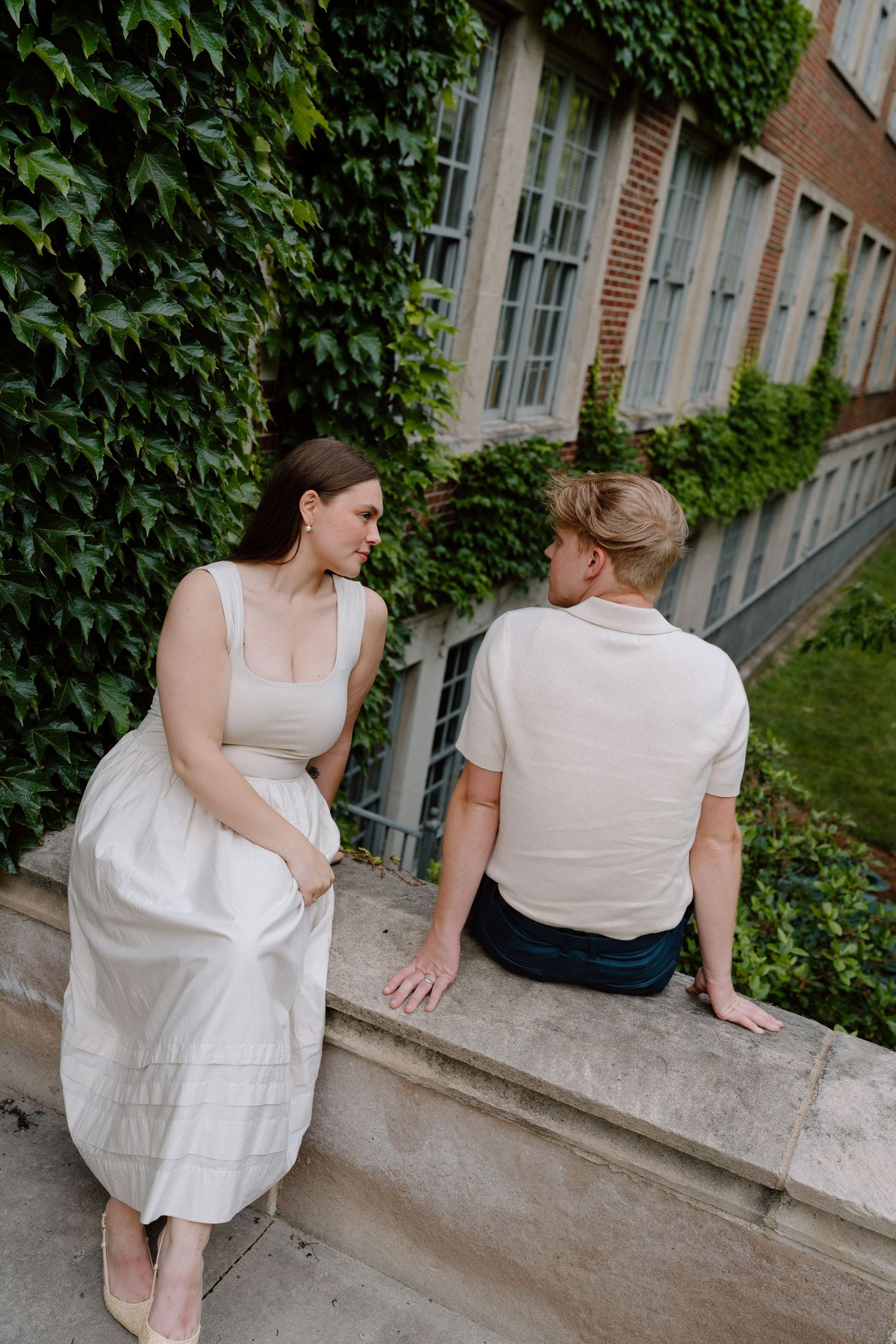 Two people sitting and talking on a stone ledge by an ivy-covered building.