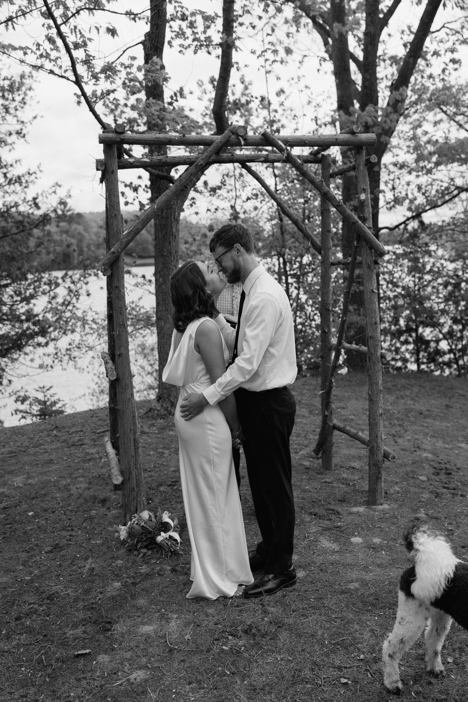 Wedding couple shares romantic moments under trees with their dog present.