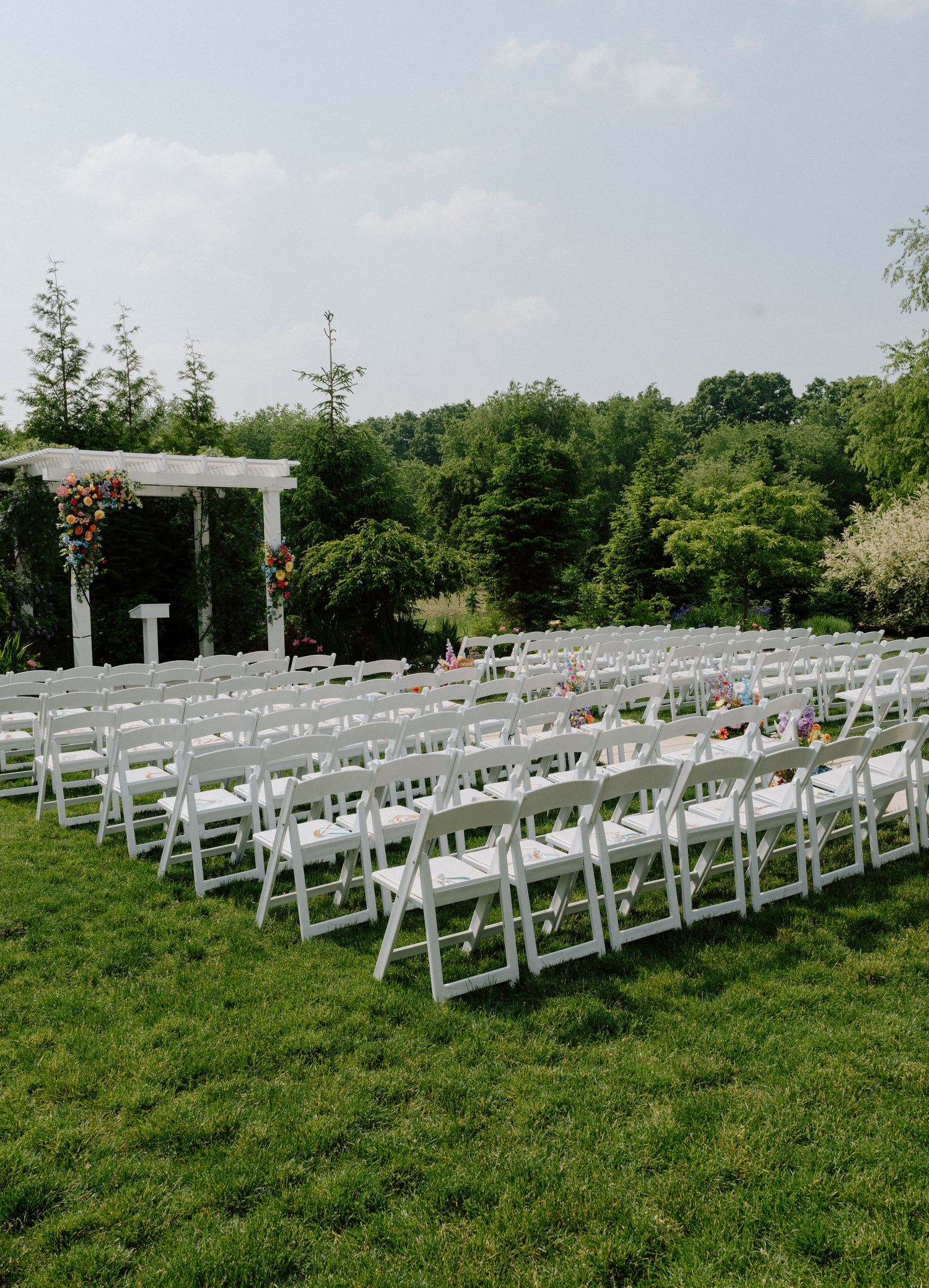 Outdoor wedding ceremony setup with rows of white chairs and floral-decorated pergola against a scenic landscape.