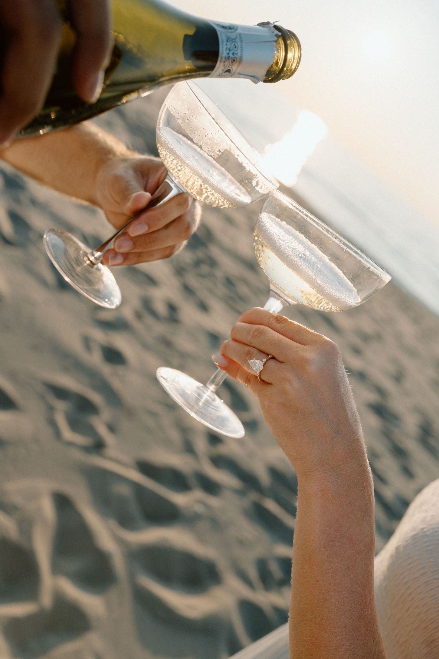 Hands pouring champagne into glasses on a beach at sunset.