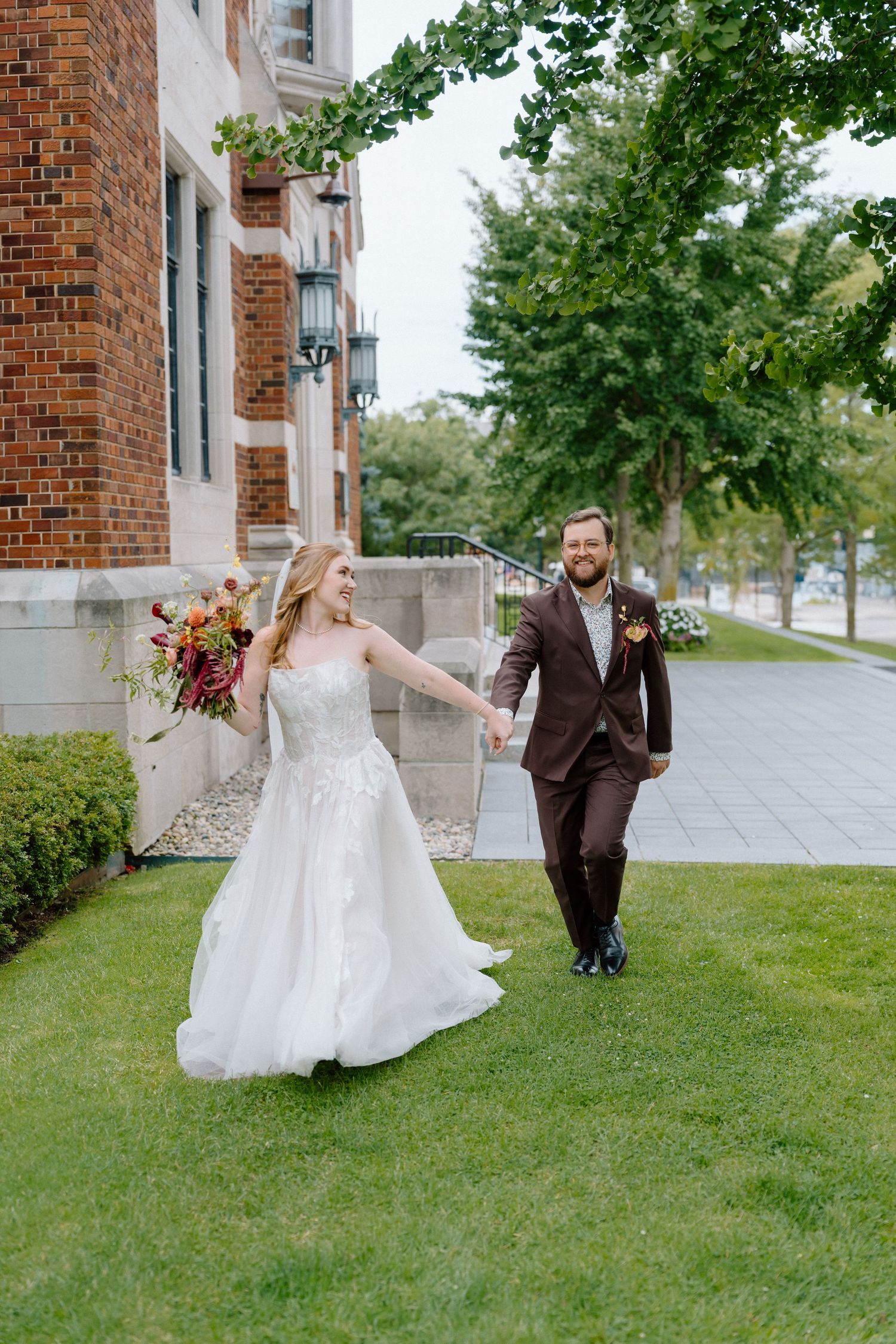A couple in wedding attire dances playfully on a green lawn outside a brick building with joyful expressions.