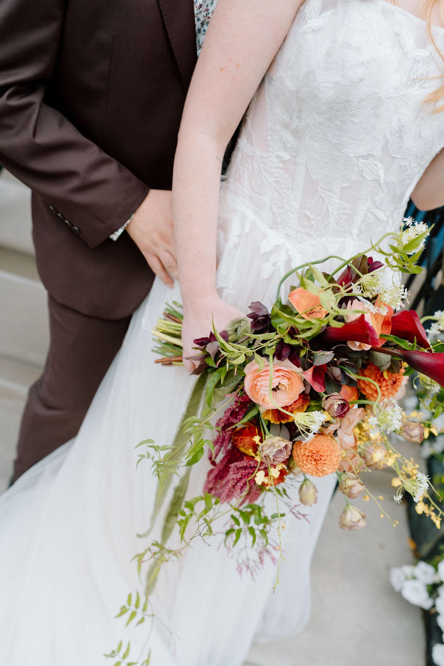 A wedding bouquet featuring vibrant orange, burgundy and yellow flowers with cascading greenery against a white dress.