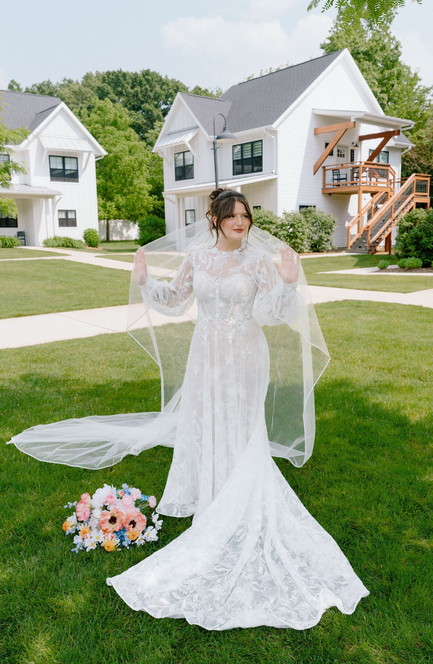 Elegant lace wedding dress with flowing sleeves displayed on green lawn with white houses in background.
