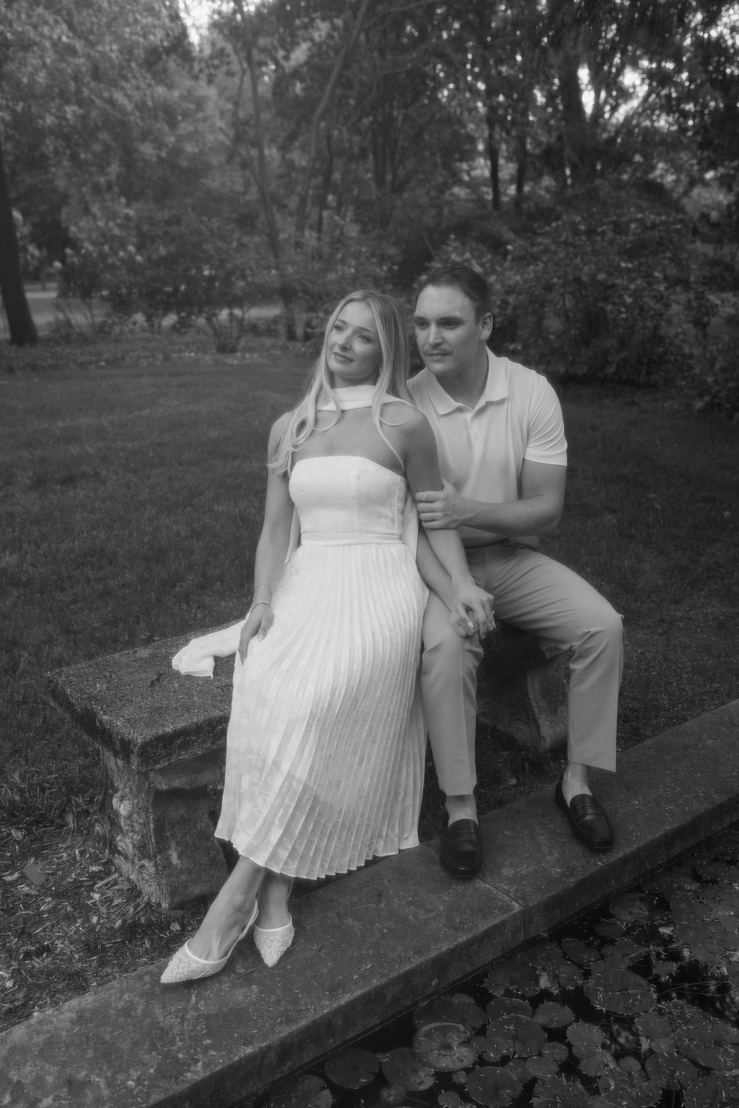 A couple sits together on a rustic wooden bench in a garden setting, captured in black and white photography.