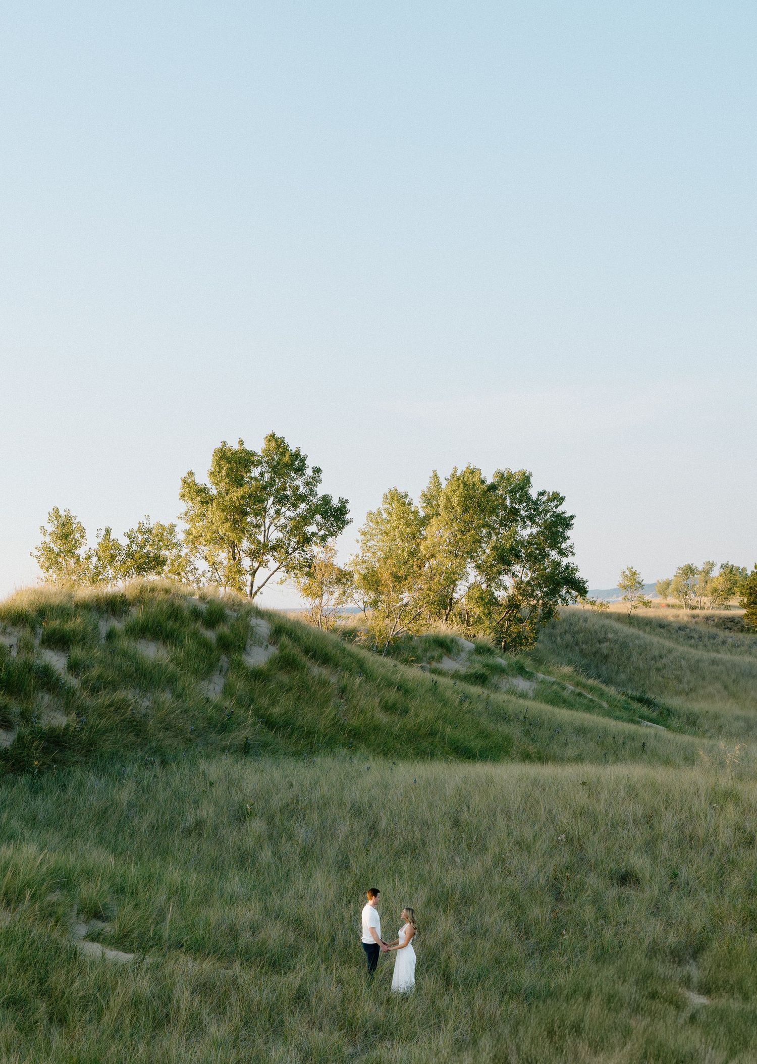 Two people stand on a grassy hillside during golden hour with trees silhouetted against the sky.