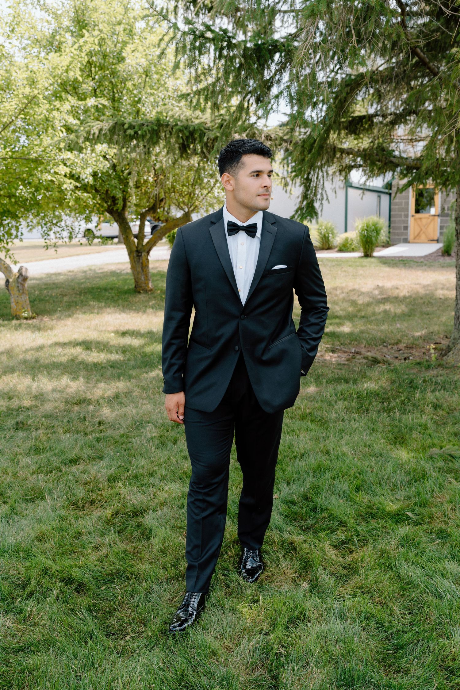 Person in black tuxedo and bowtie stands on grassy hillside near evergreen trees.