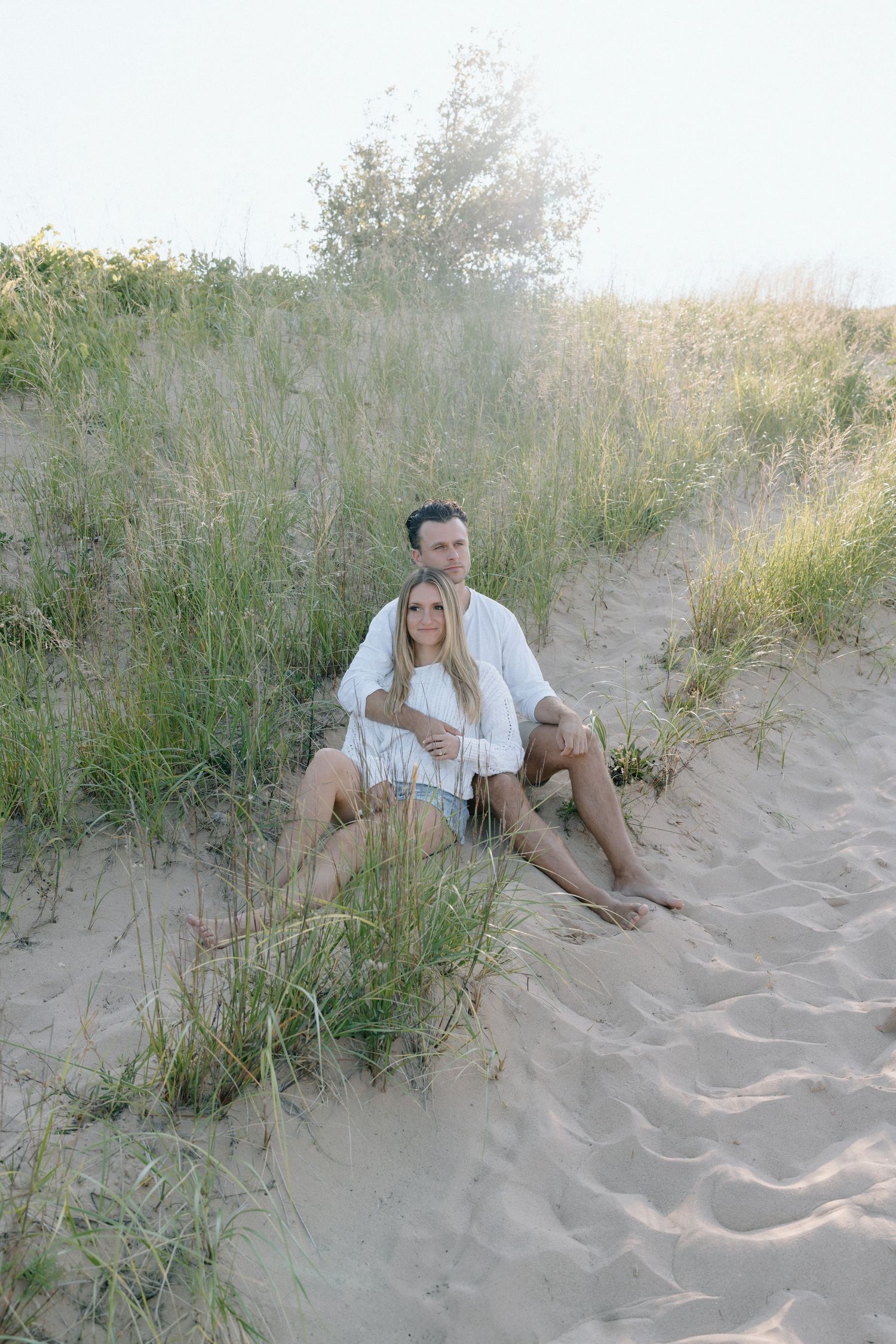 Couple sitting together on sandy dunes surrounded by beach grass at sunset.