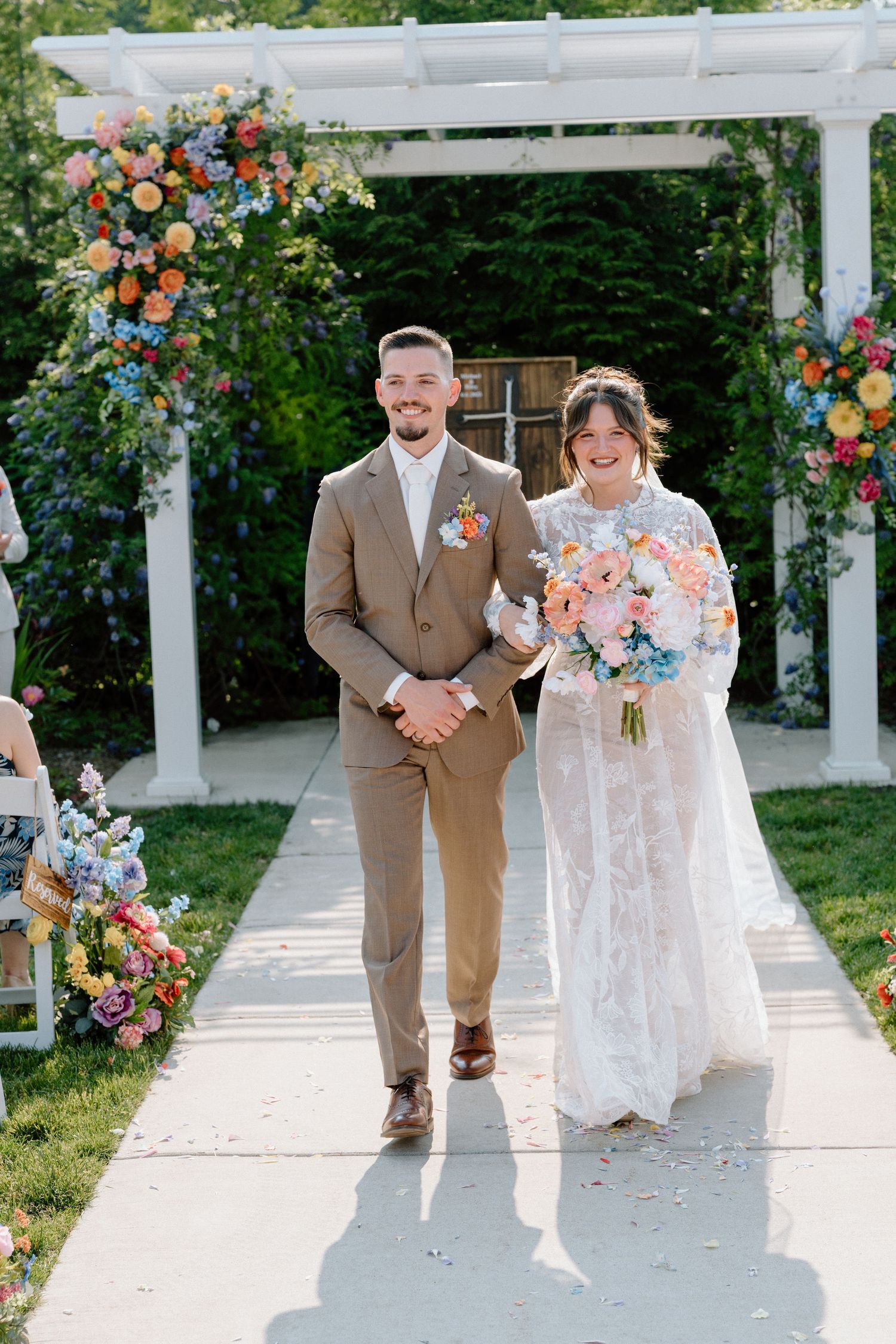 Couple walks along a white pergola decorated with vibrant coral and peach floral arrangements.