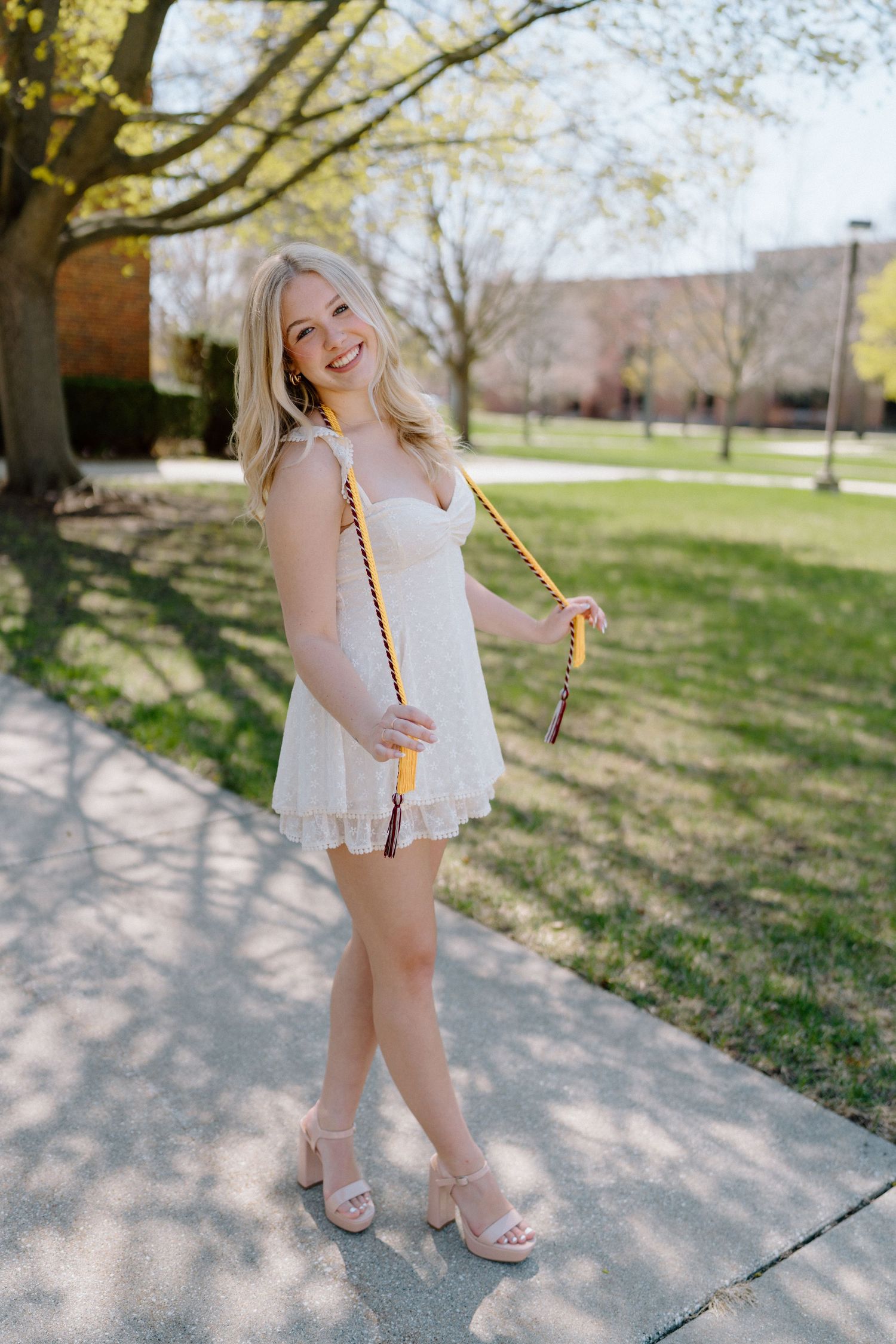 Graduate in white dress with gold tassel poses outdoors on sunny spring day.