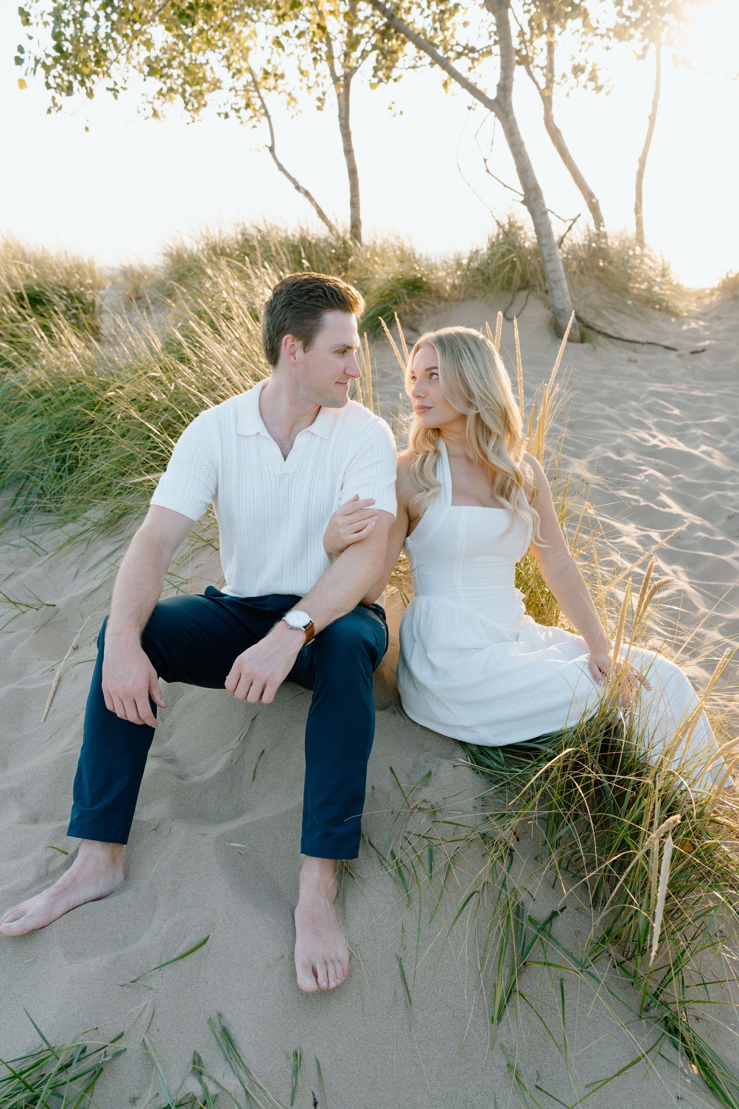 Couple sitting on sandy beach dunes at sunset with tall grass swaying in the golden light.