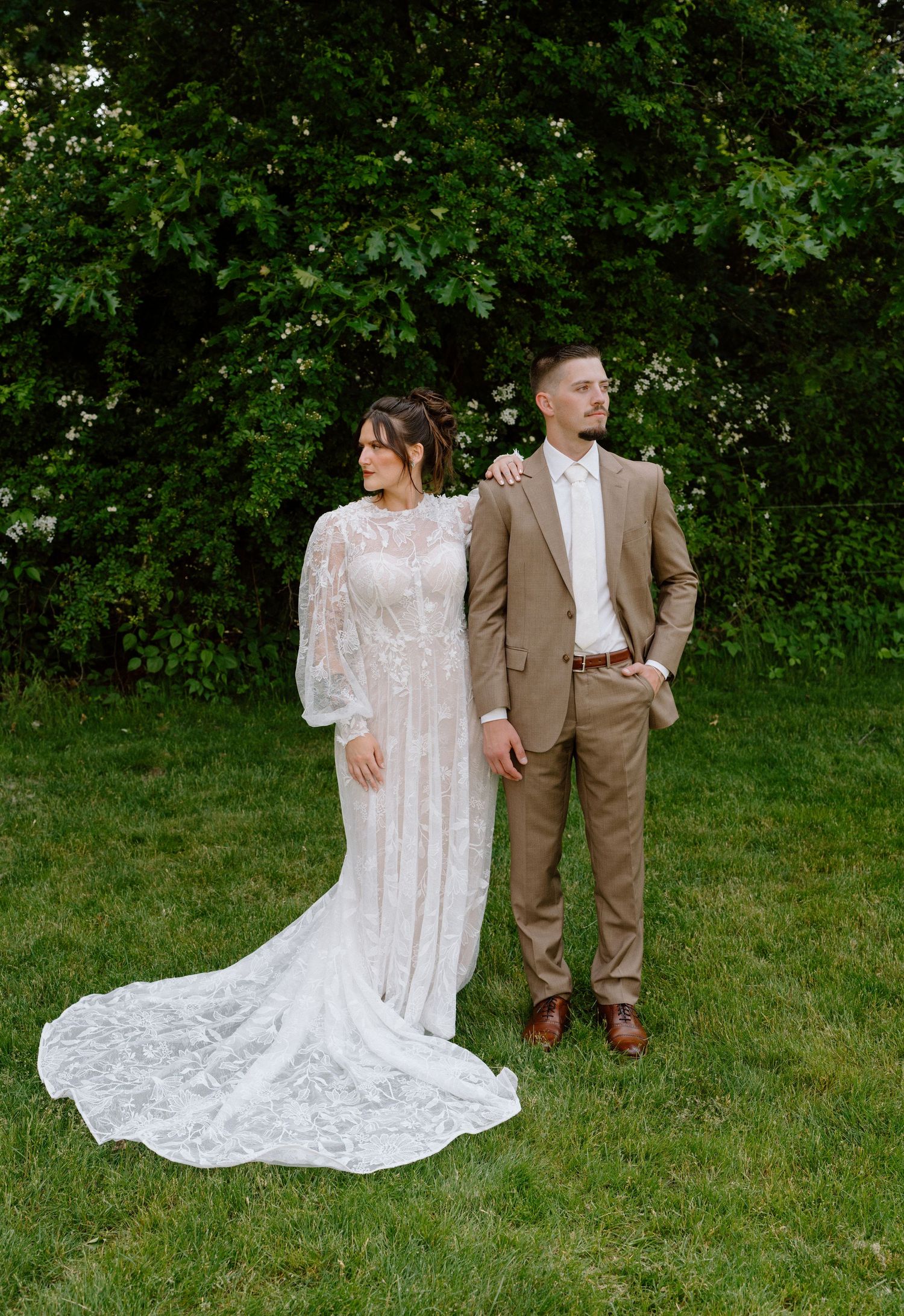 Wedding couple poses on green lawn with bride wearing flowing lace dress and groom in brown suit.