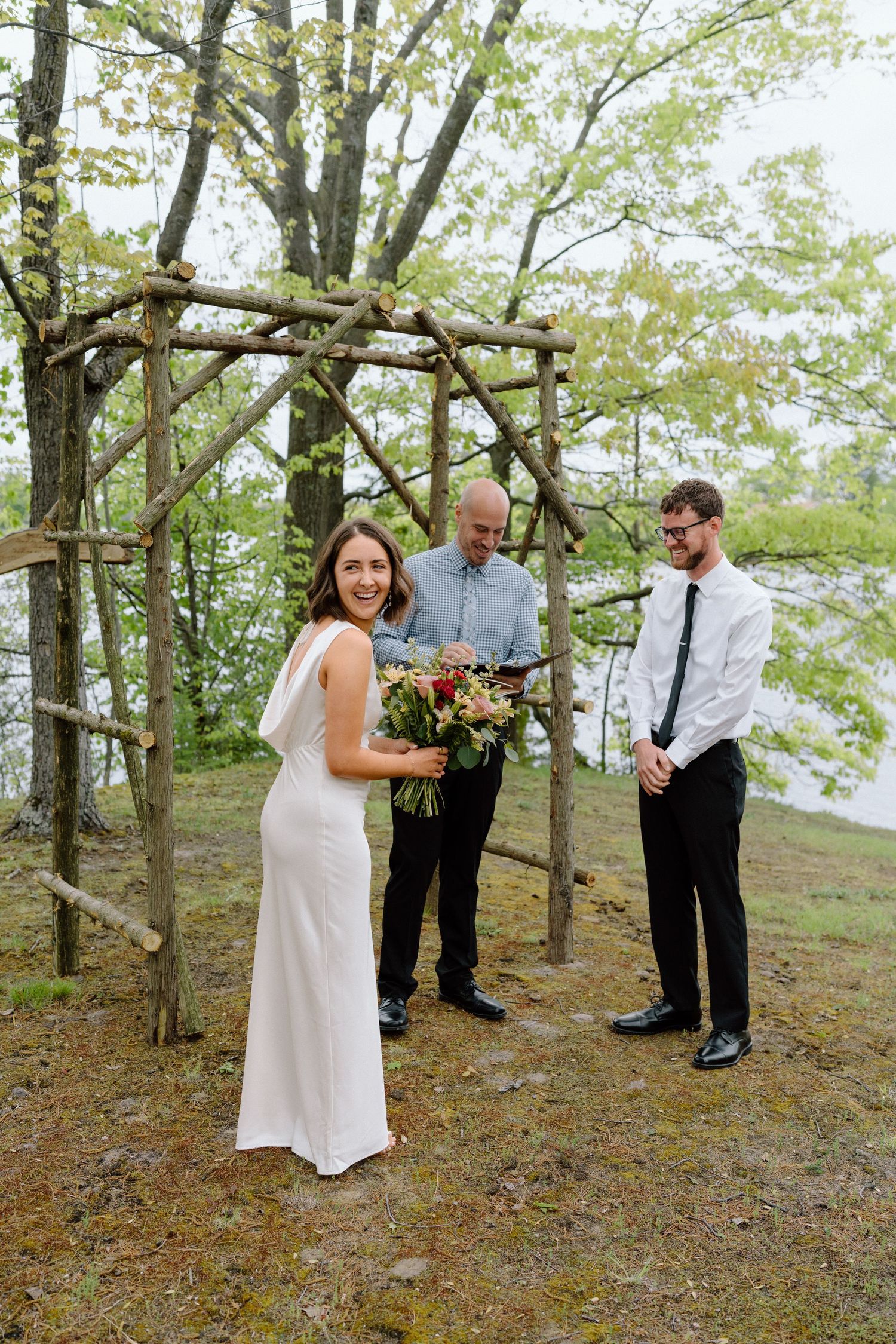 Wedding ceremony participants in white and formal wear gather beneath trees in a wooded setting.