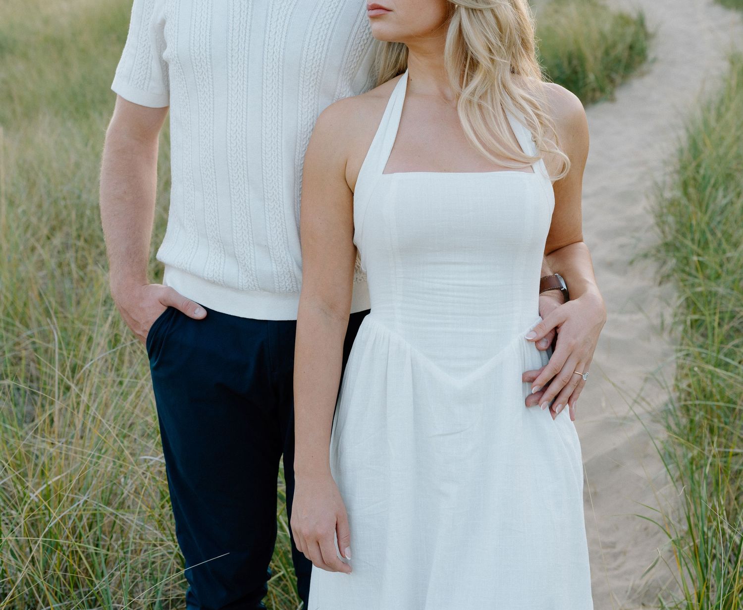A couple in wedding attire walking together on a dirt path through a grassy field.