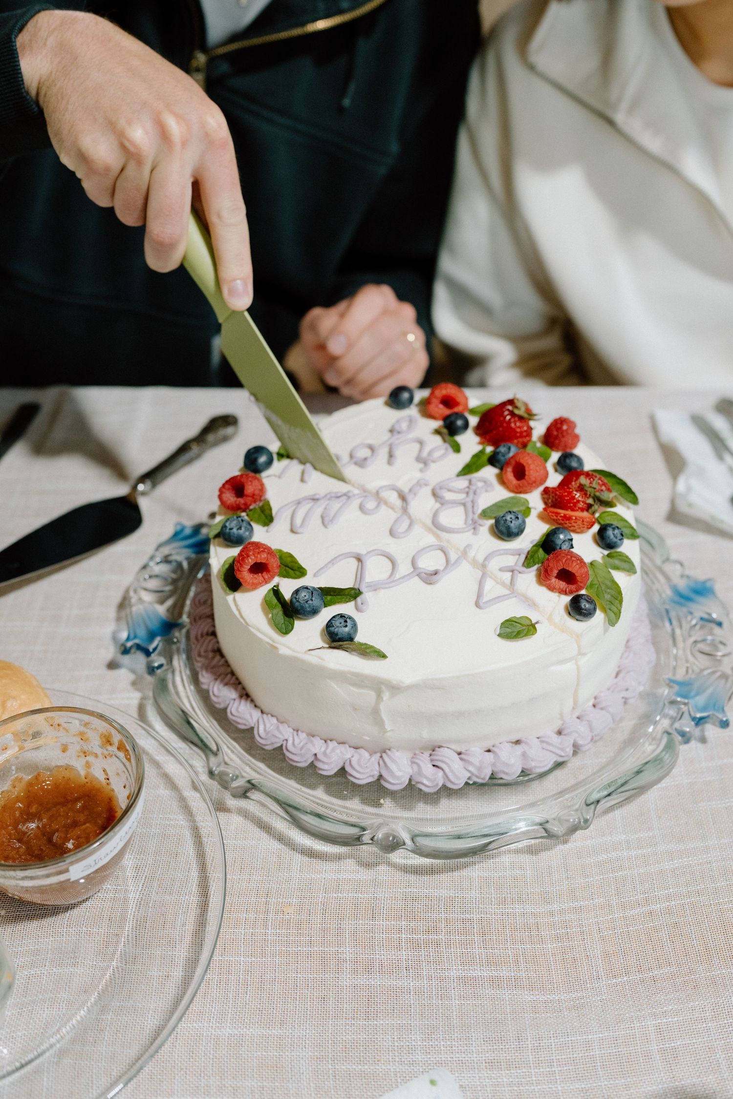 White wedding cake decorated with fresh berries and flowers being cut on a lace tablecloth.