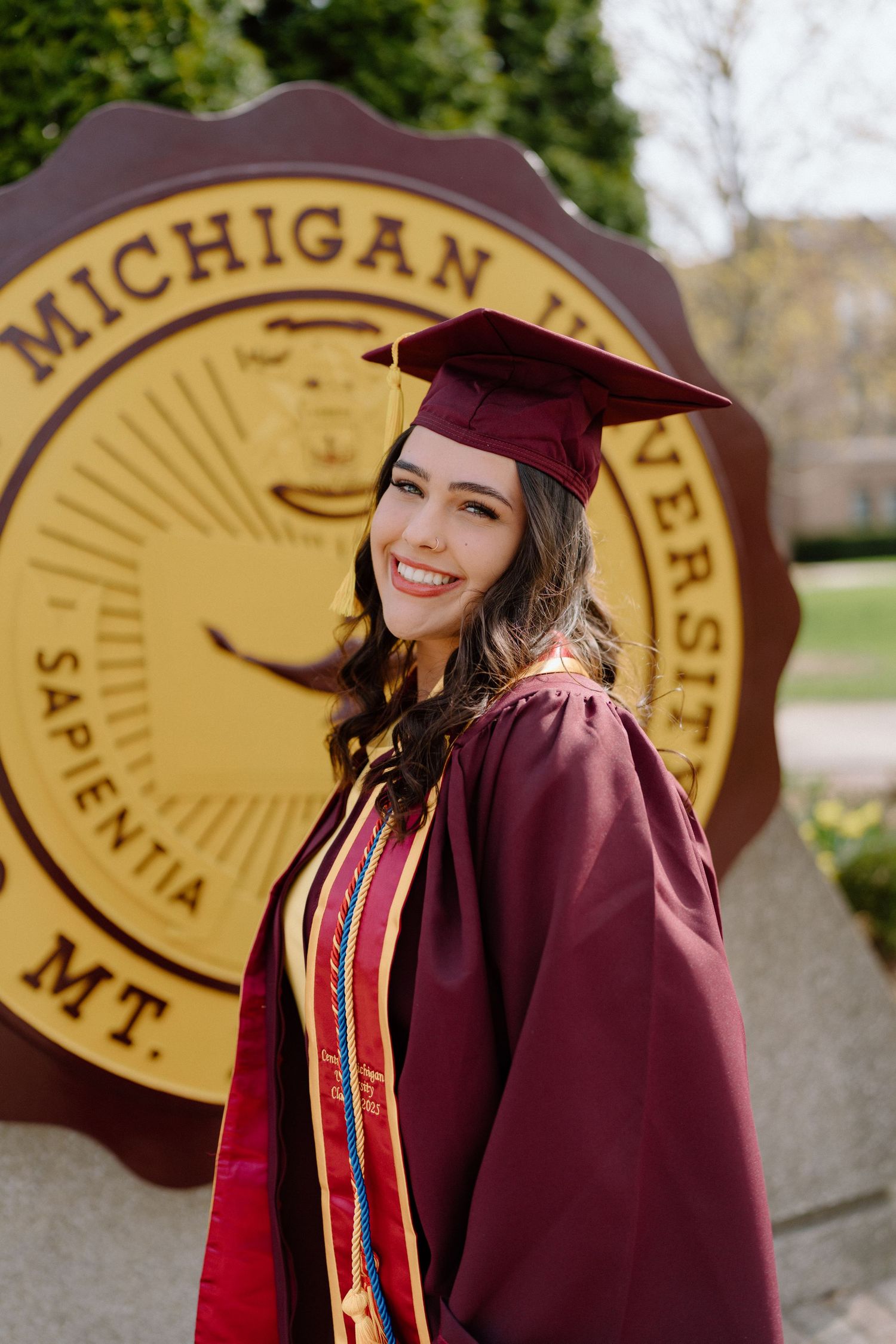 Graduation photos with maroon cap and gown in front of large golden university seal medallions.