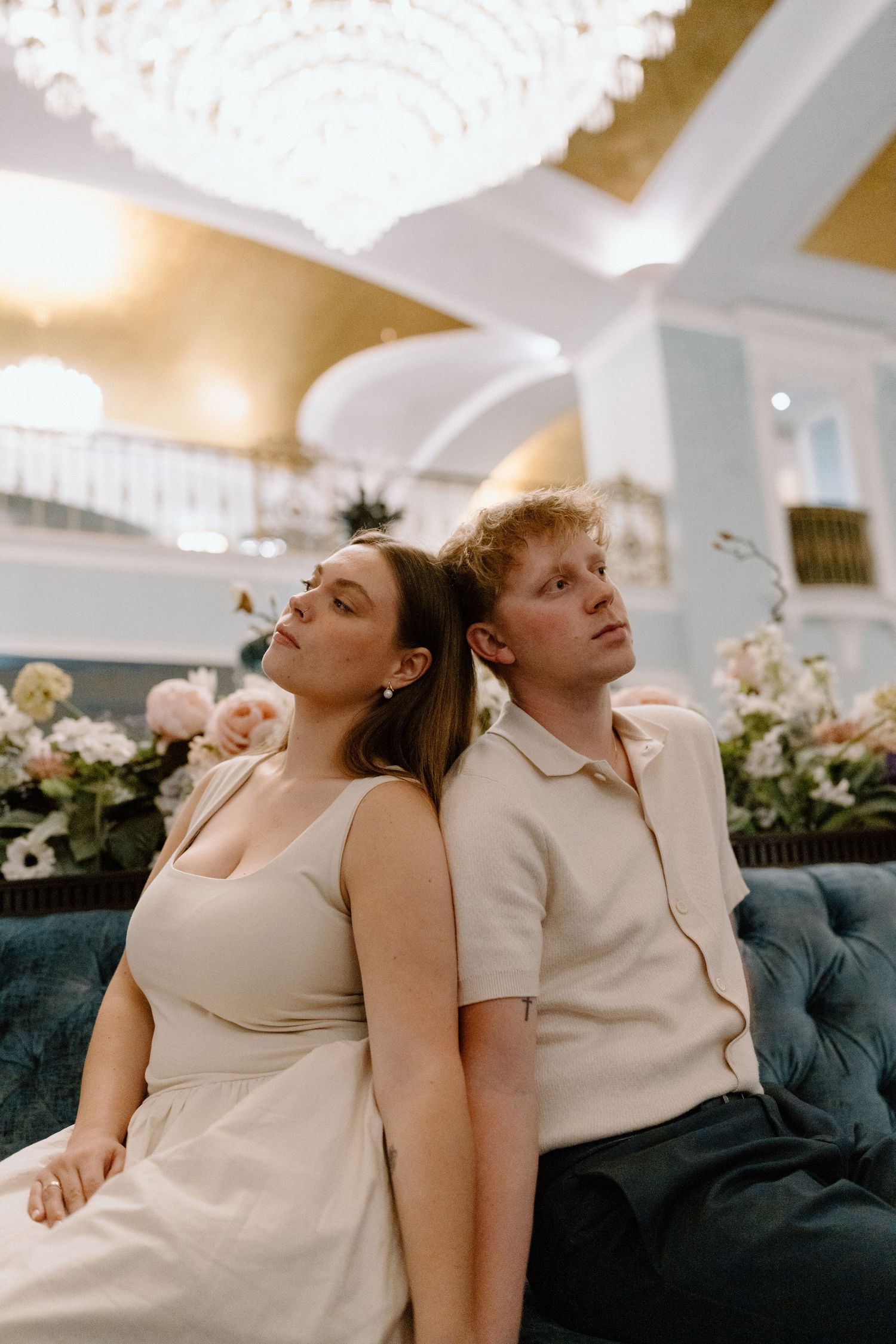 Two people relaxing on blue velvet couch under elegant chandelier with floral arrangements.