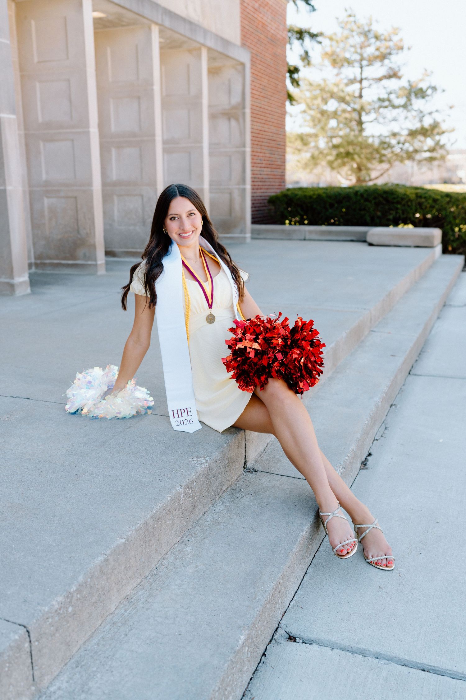 Young woman in graduation attire holding red and white pom-poms, seated on steps outdoors, wearing a medal and sash.