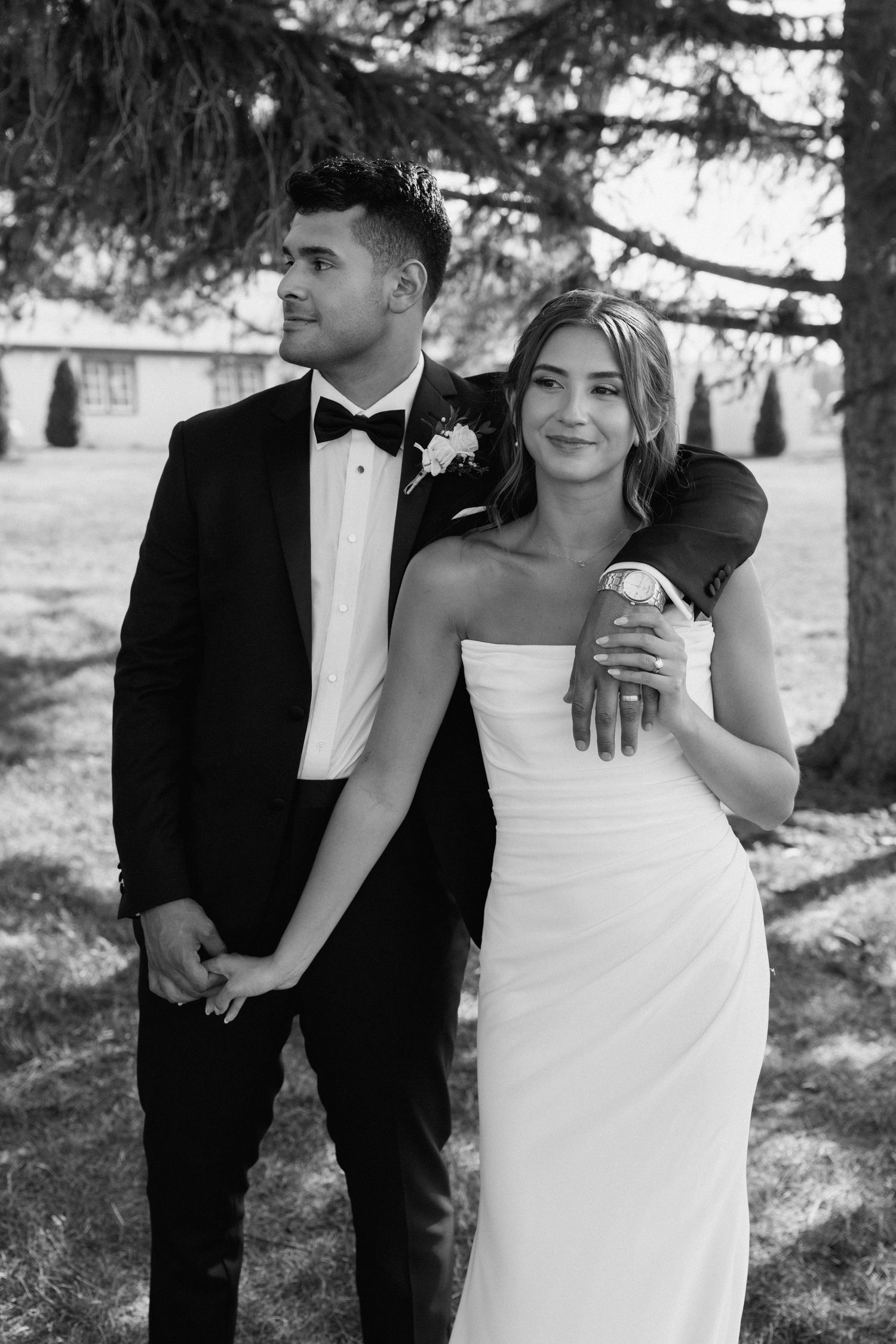 Wedding couple poses together in formal attire under evergreen trees in black and white.