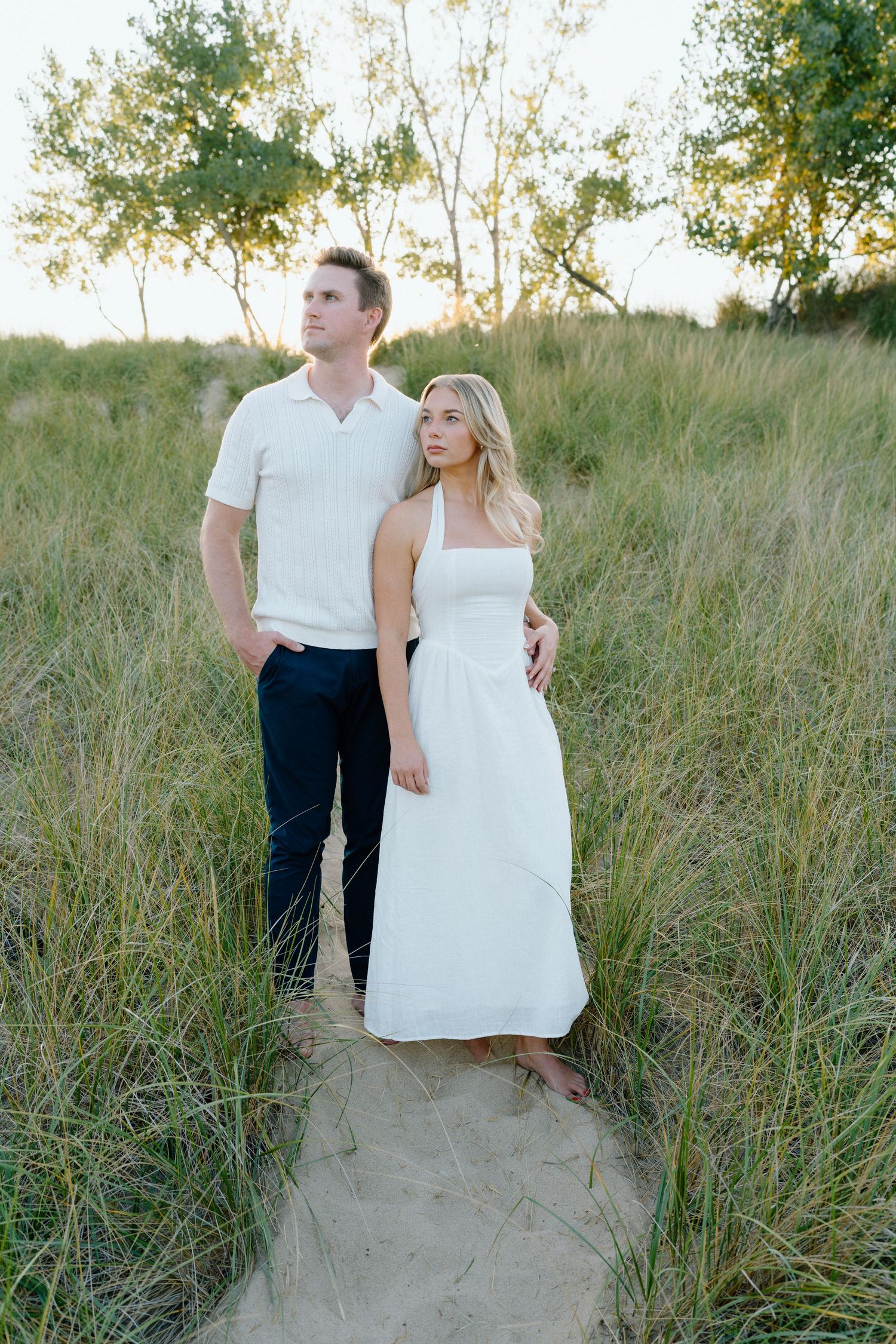 A couple stands together in tall grass and sand dunes during a sunset photoshoot wearing white outfits.