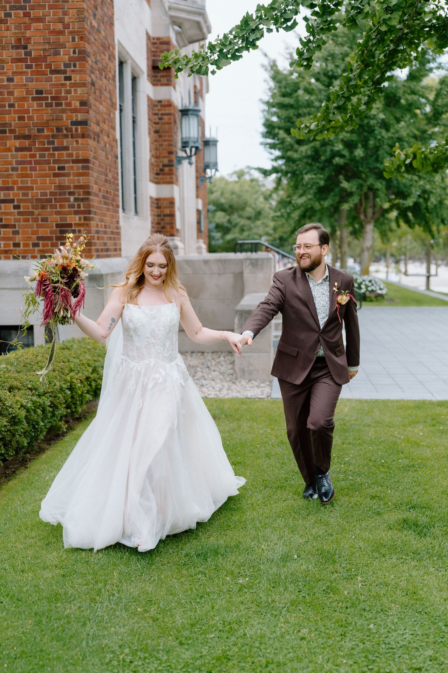 Couple in wedding attire dances and spins playfully on a grassy lawn in front of a brick building.