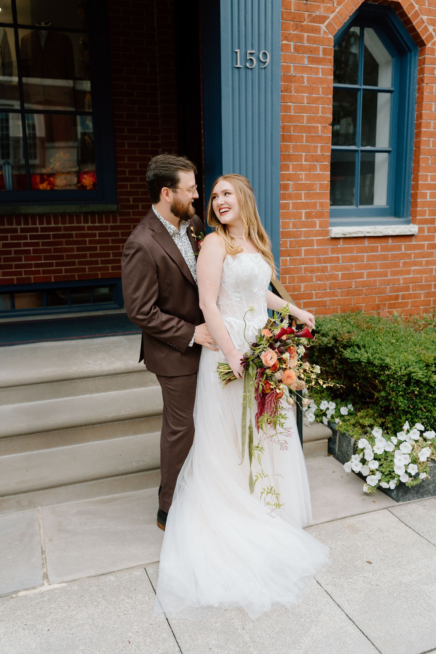A wedding couple shares a joyful moment outside a brick building with blue trim details.