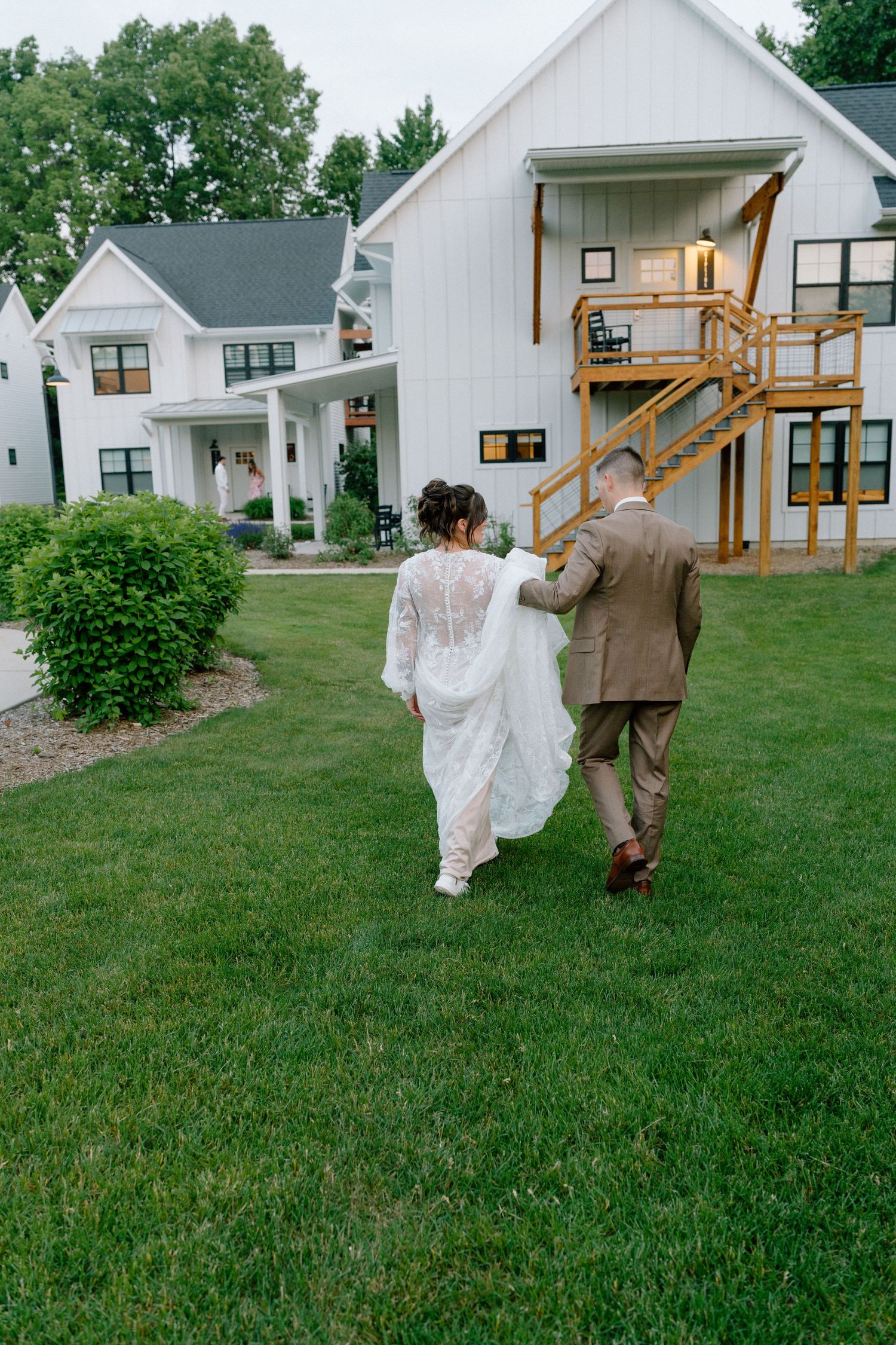 Couple walking across lawn towards white farmhouse buildings in wedding attire.