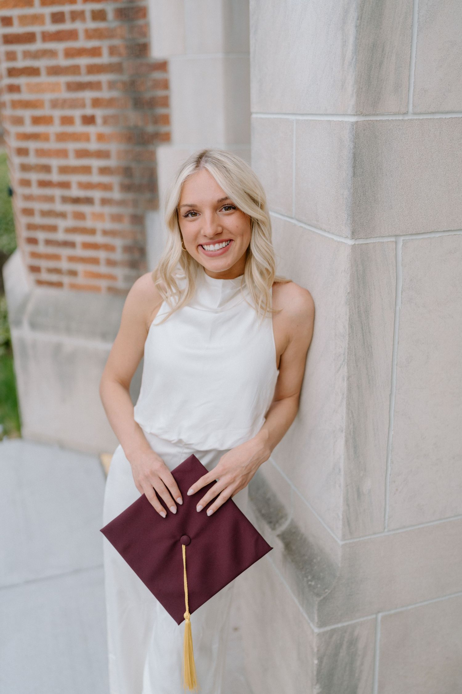 Graduate holds burgundy diploma cover while posing against stone building wall.
