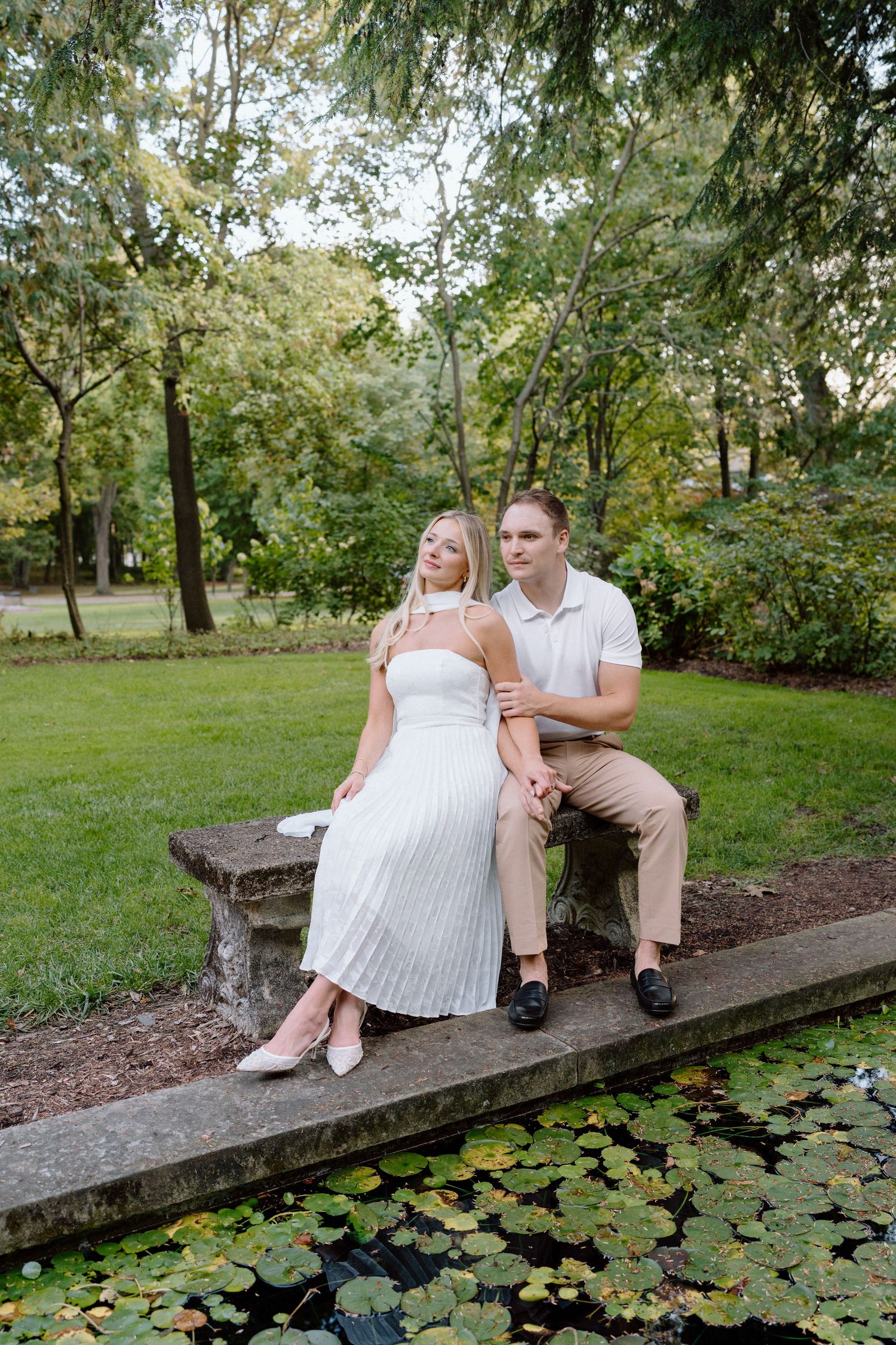 Couple sits together on concrete ledge beside tranquil pond with lily pads in lush garden setting.