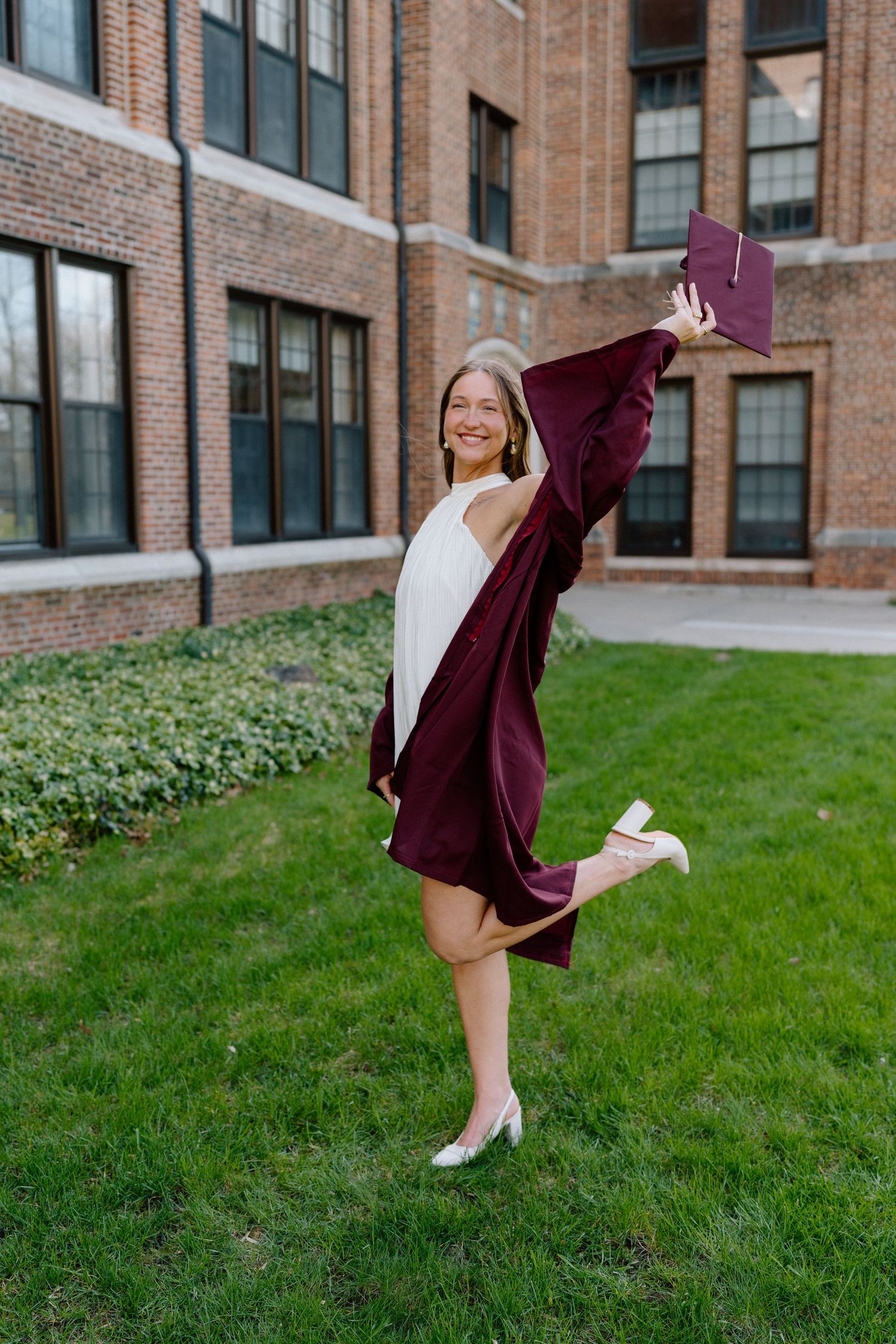 Smiling woman in maroon graduation gown holding diploma, posing joyfully on green lawn outside brick university building.