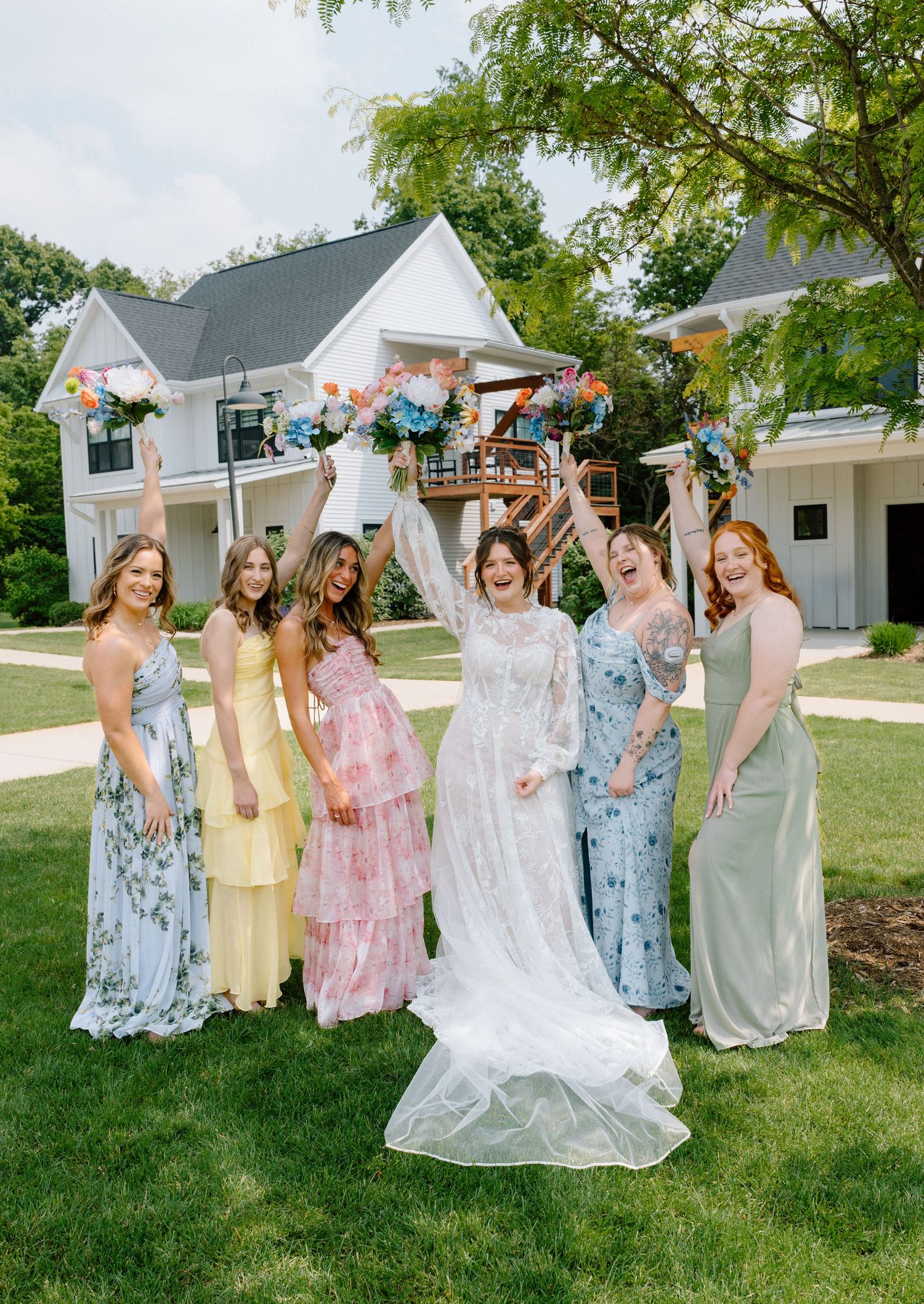 Bridal party in pastel and floral dresses poses joyfully on a sunny lawn.