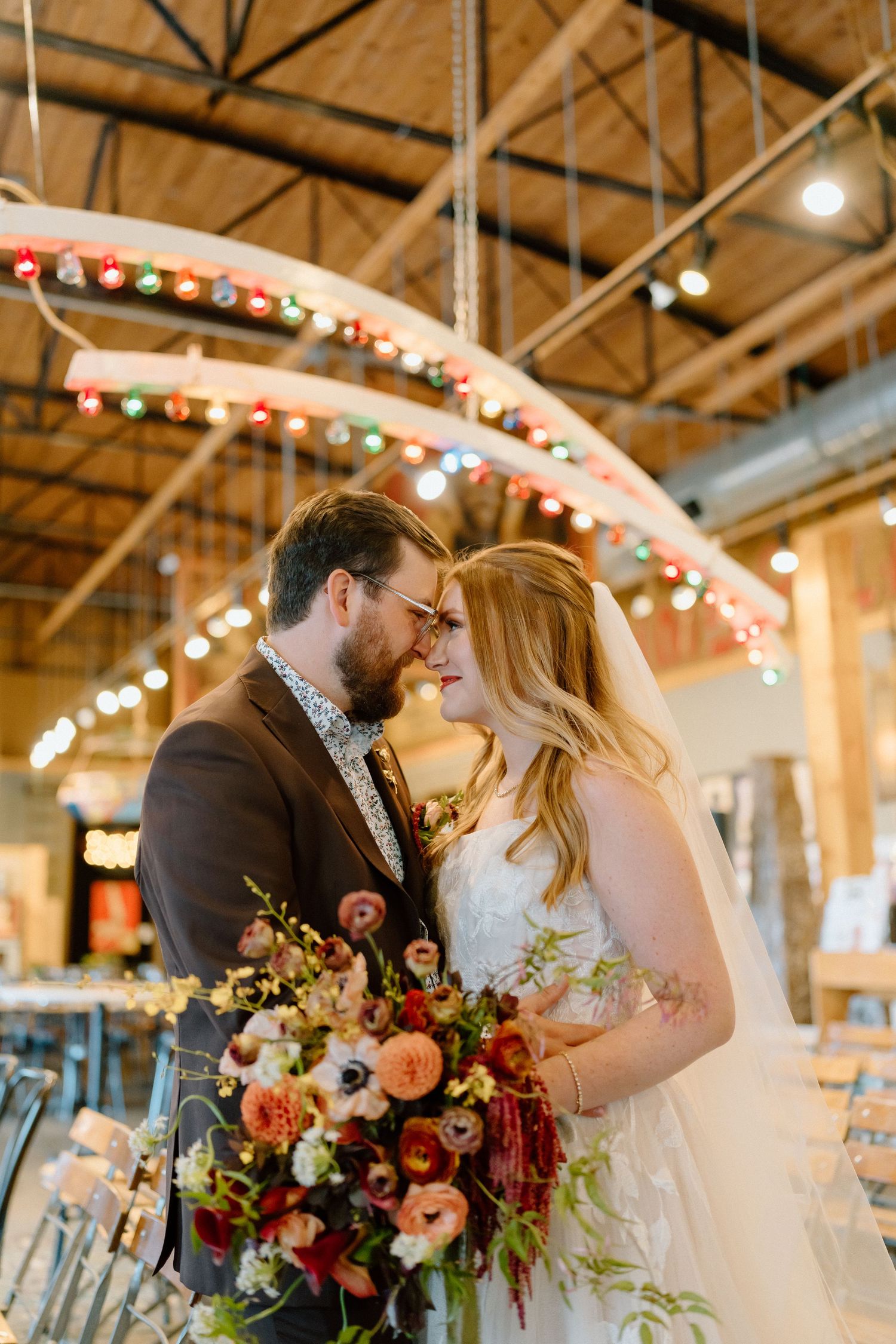 Romantic wedding couple shares intimate moments in rustic industrial venue decorated with string lights and floral arrangements.