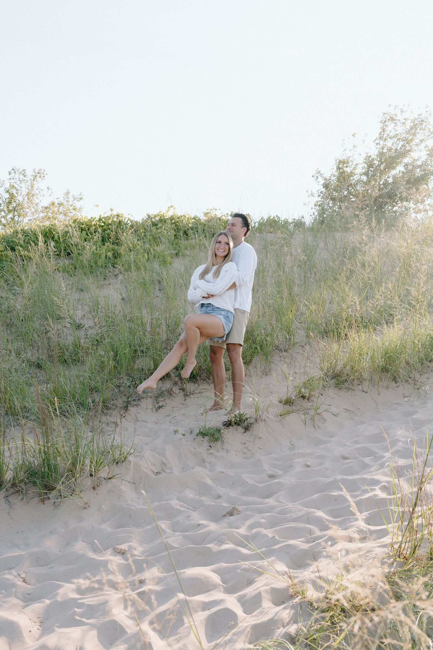 Two people posing together on sandy beach dunes with coastal vegetation.