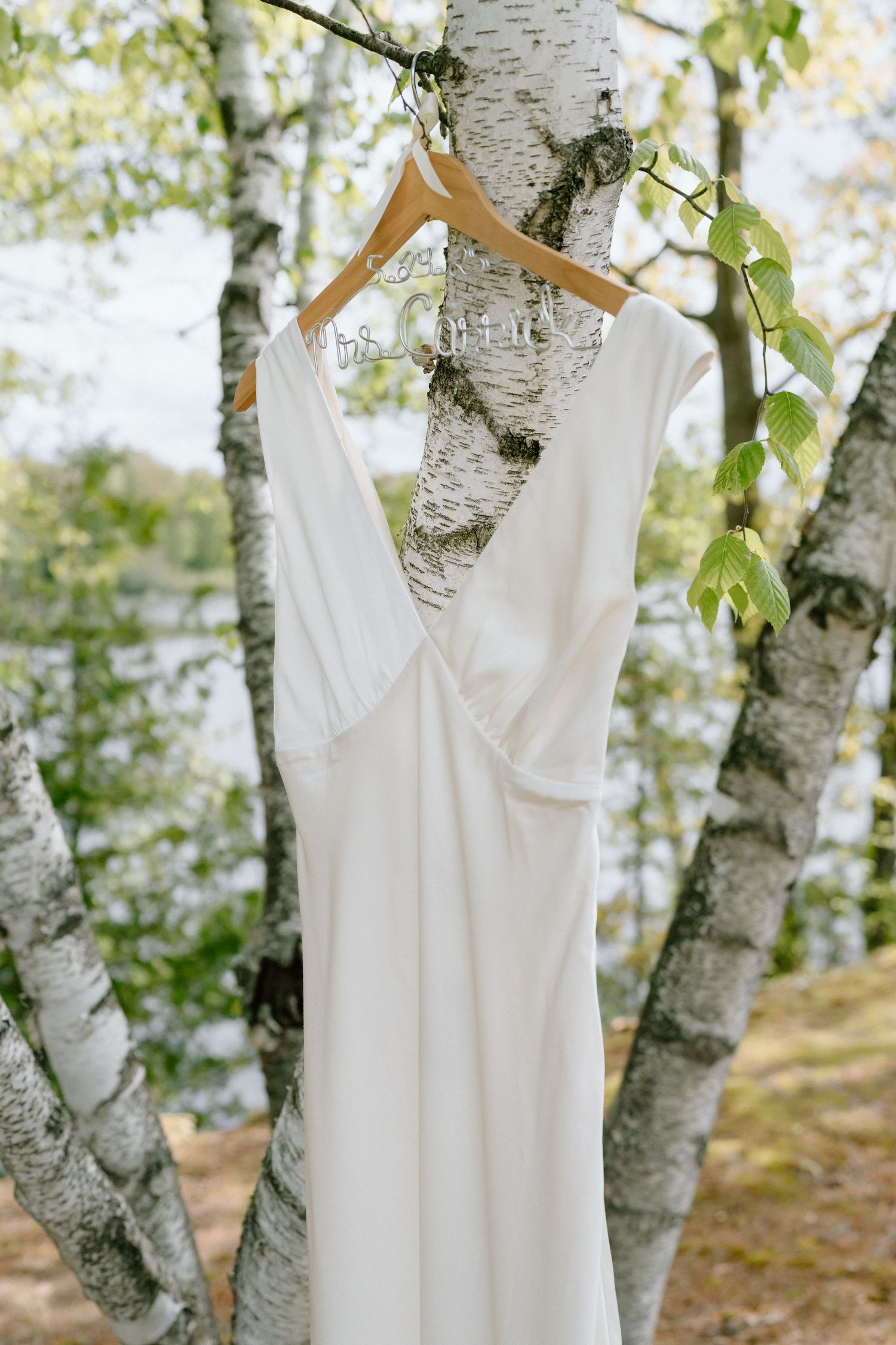 Elegant white wedding dress hanging on wooden hanger against birch trees in natural setting.